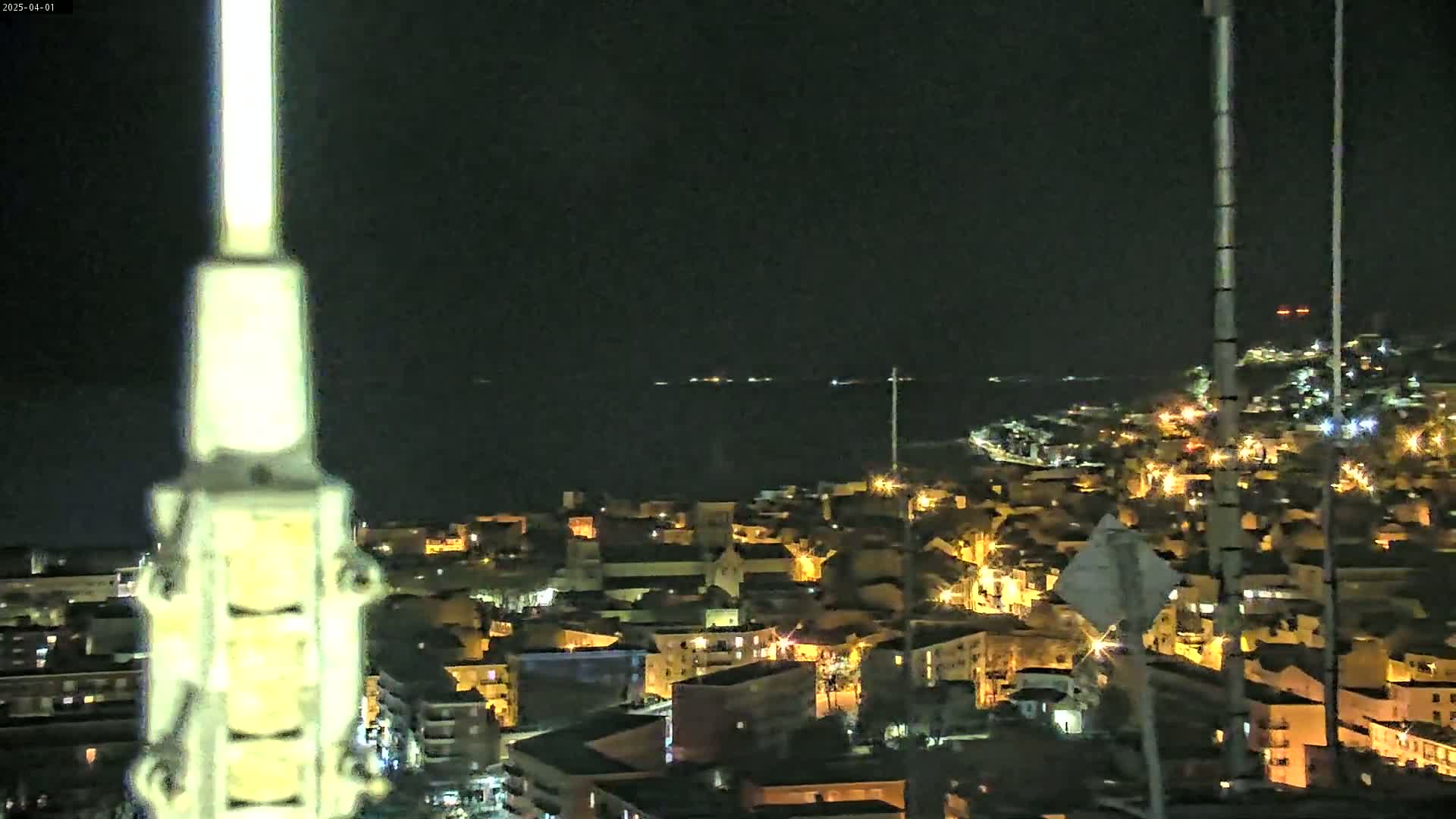 A nighttime, high-angle view shows a coastal town illuminated by streetlights, with a dark body of water visible in the background under a clear sky.