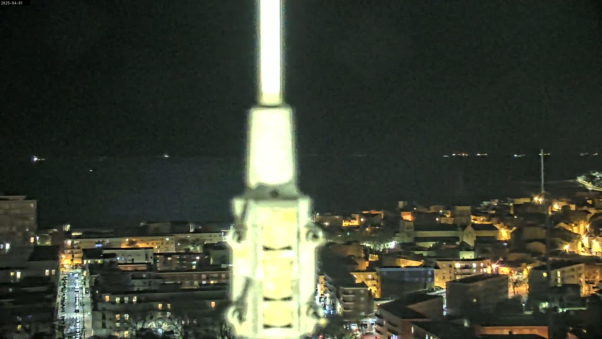 A brightly lit tower stands in the foreground, overlooking a dark, nighttime cityscape with a calm body of water visible in the distance.
