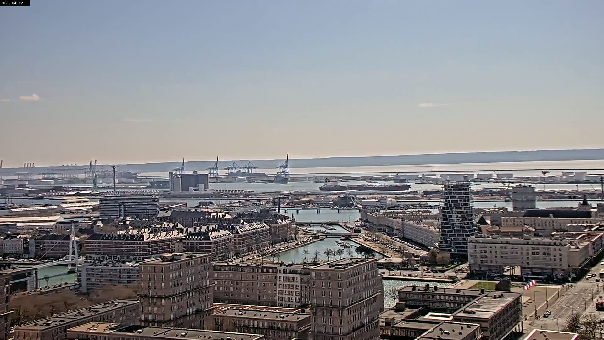 A sunny day overlooking a harbor city with numerous buildings, a canal, and a large industrial port in the distance.