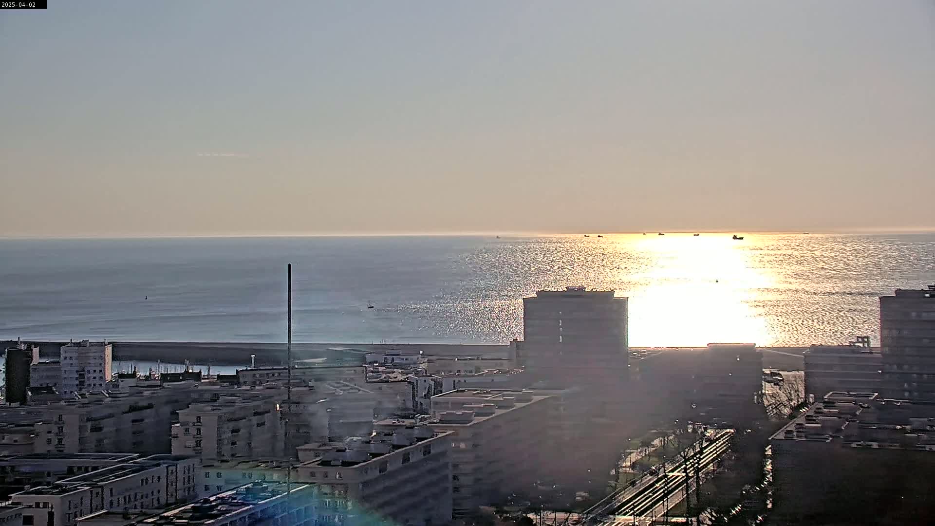 A city skyline with several multi-story buildings is seen overlooking a calm ocean reflecting the setting sun on a clear day.
