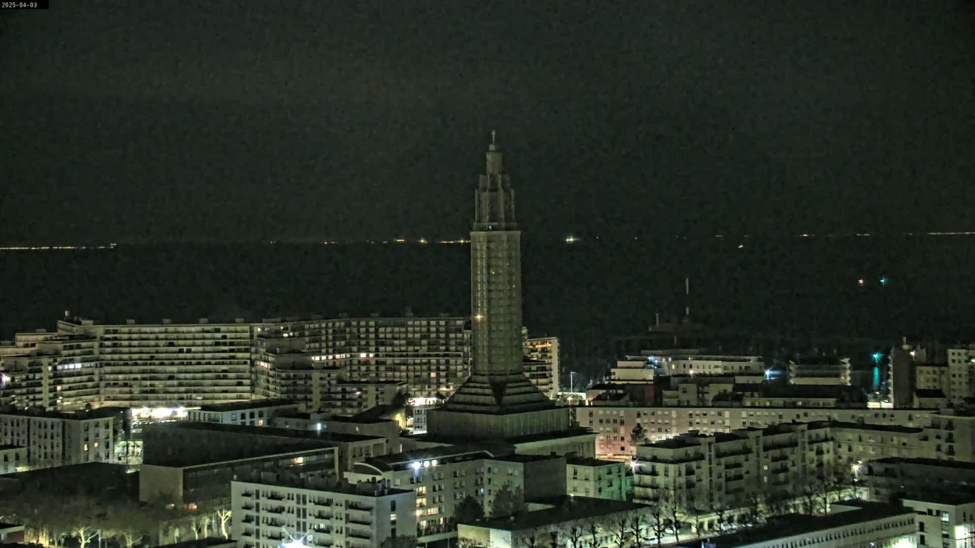 A tall, slender tower stands illuminated in a nighttime cityscape overlooking a dark body of water under a clear sky.