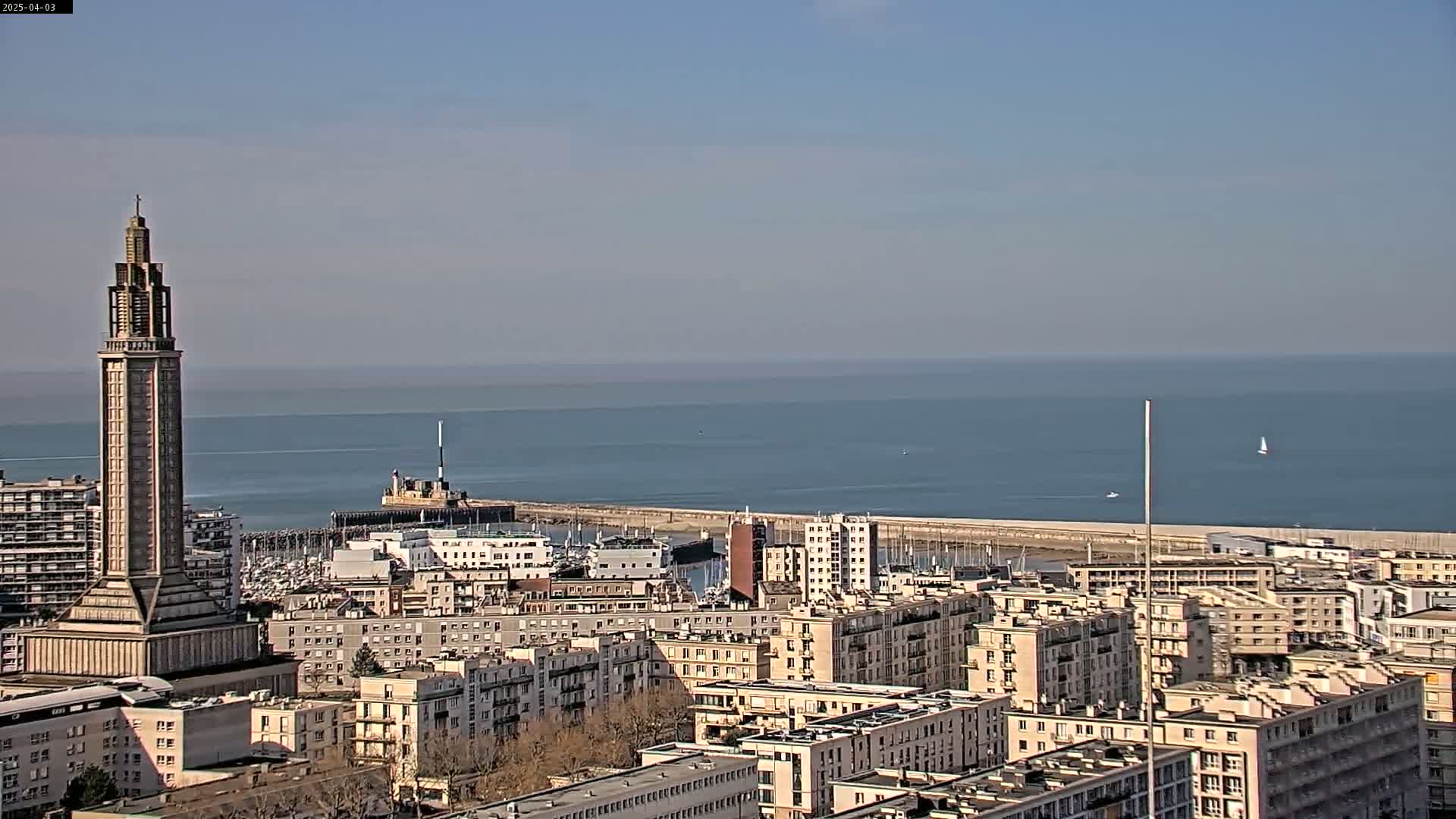 A tall, slender tower stands prominently in a city overlooking a calm ocean under a clear sky.