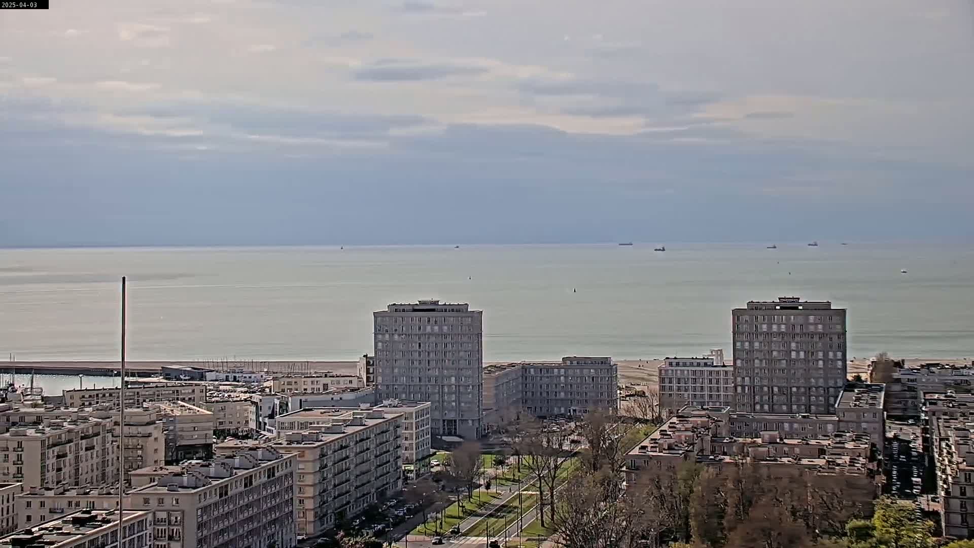 A coastal city with several multi-story buildings is seen under a mostly cloudy sky, with a calm sea and a few ships visible in the distance.