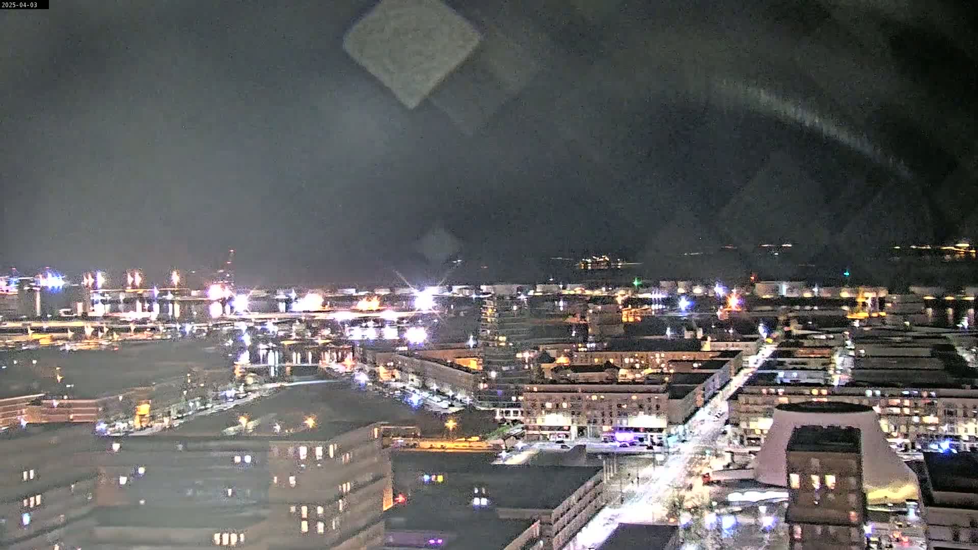 A nighttime aerial view of a city with illuminated buildings and streets, under a dark, cloudy sky.
