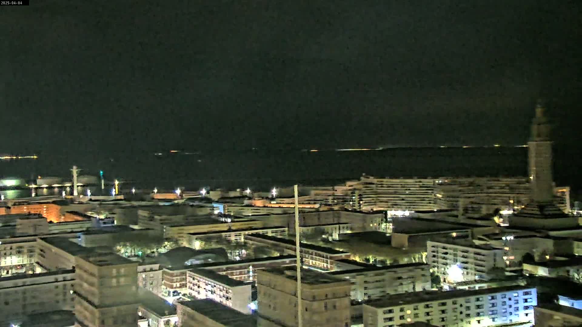 A nighttime aerial view shows a city illuminated near a dark body of water under a clear, dark sky.