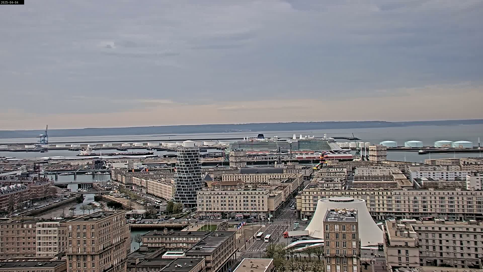A large cruise ship is docked at a harbor in a city on an overcast day, with numerous buildings and industrial structures visible.