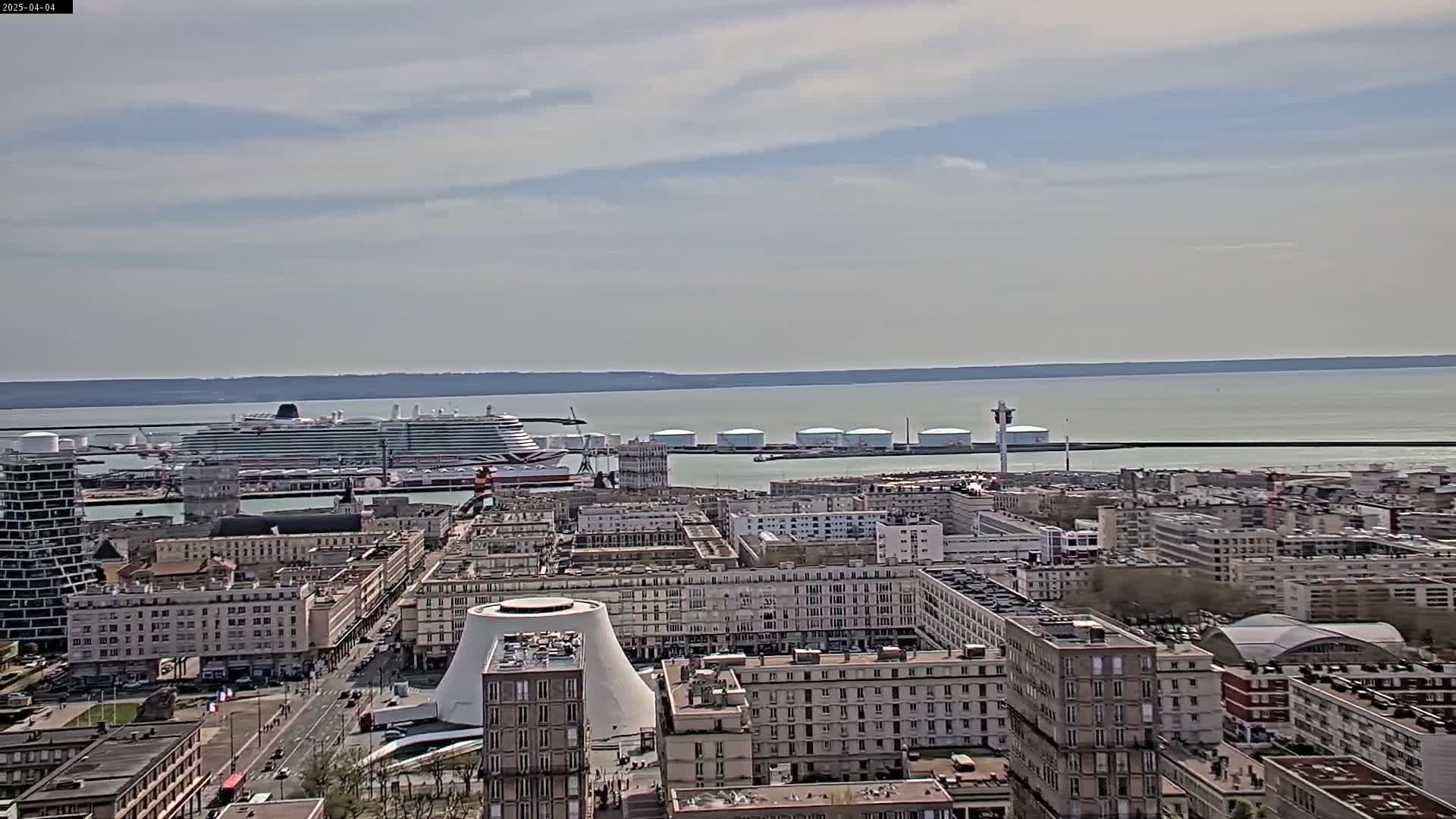 A large cruise ship is docked at a port city under a partly cloudy sky, with a calm body of water visible in the background.