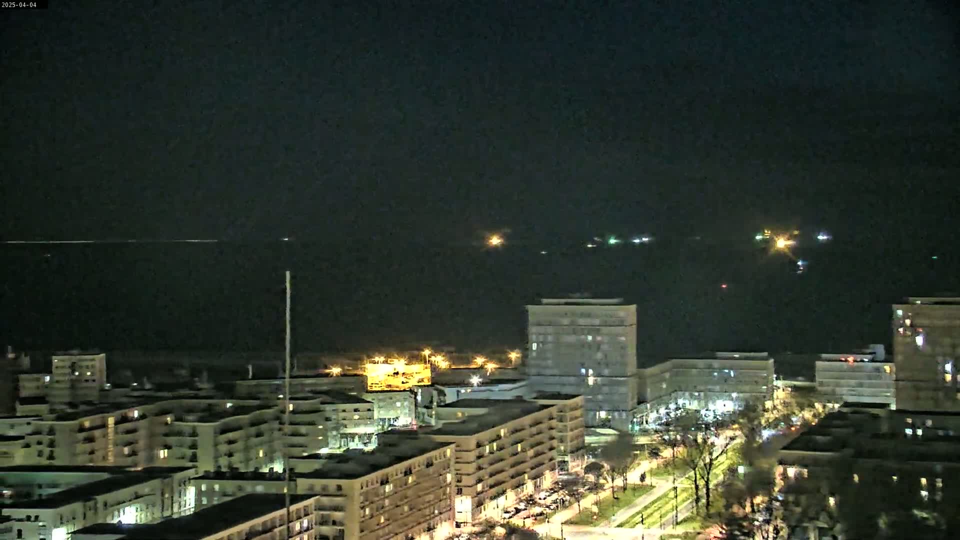 A nighttime view of a city skyline with apartment buildings and a dark body of water in the background, showing several distant lights on the water.