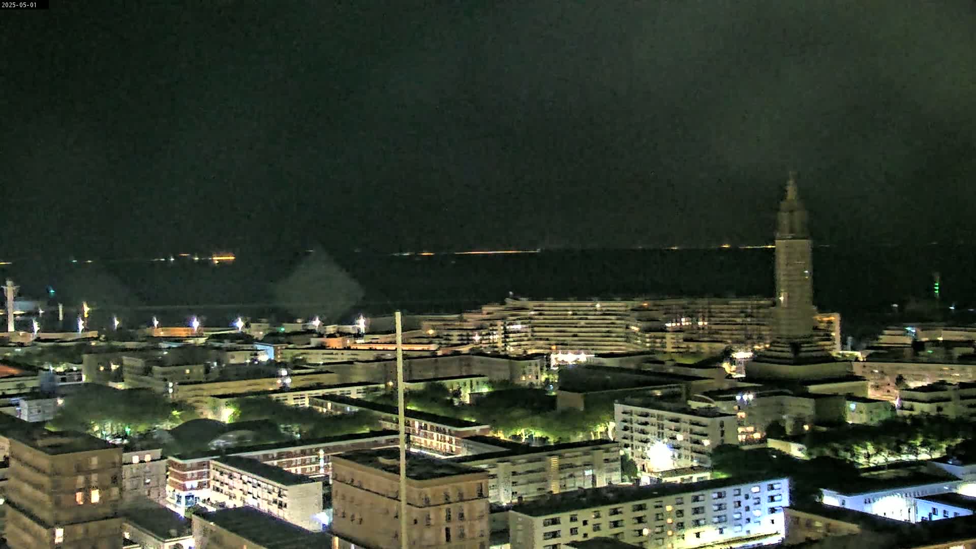 A nighttime aerial view of a city by a body of water, showing a mix of buildings with lights on, and a tall tower in the distance.