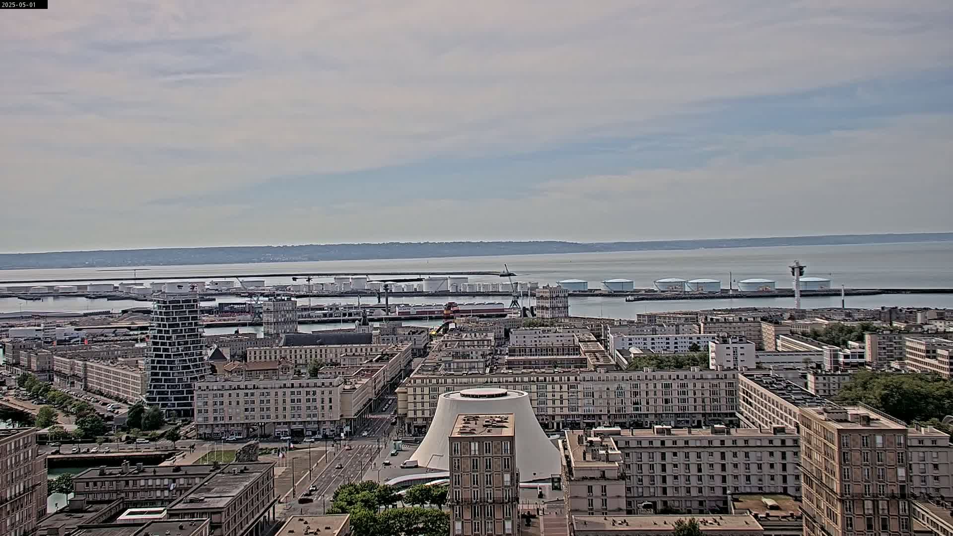 A city harbor with many buildings and industrial structures is visible, under a mostly cloudy sky.