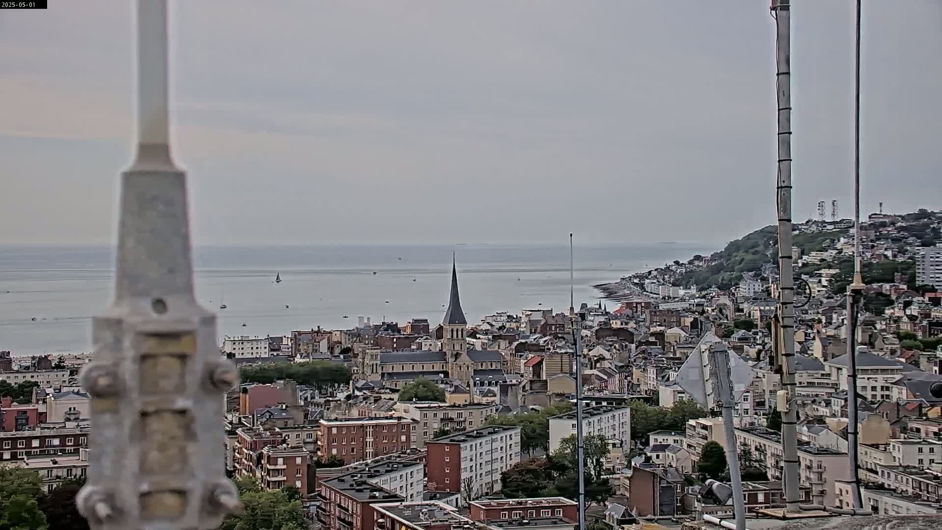 A coastal town with a church steeple is seen from an elevated vantage point on an overcast day, with a calm sea visible in the background.
