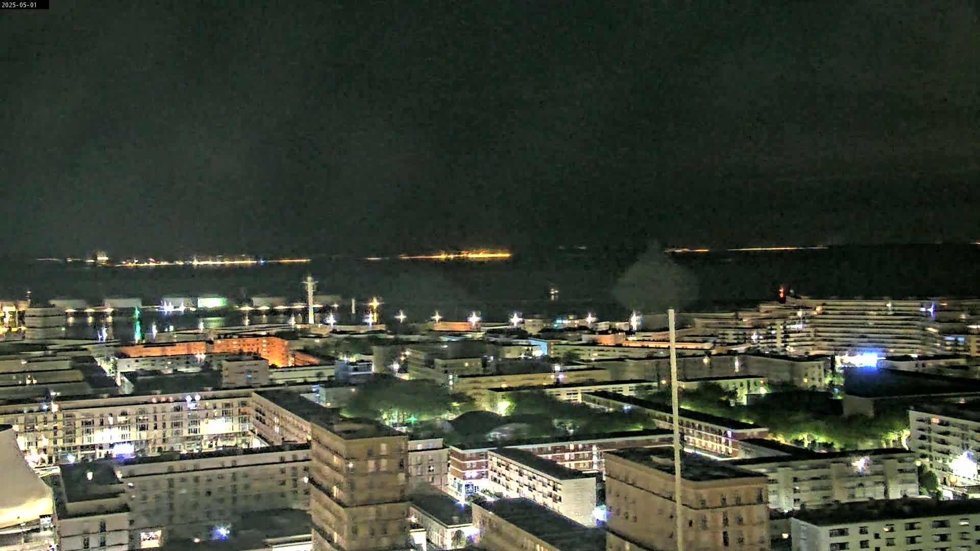 A nighttime aerial view shows a city with illuminated buildings and a dark body of water with lights visible in the distance under a clear, dark sky.