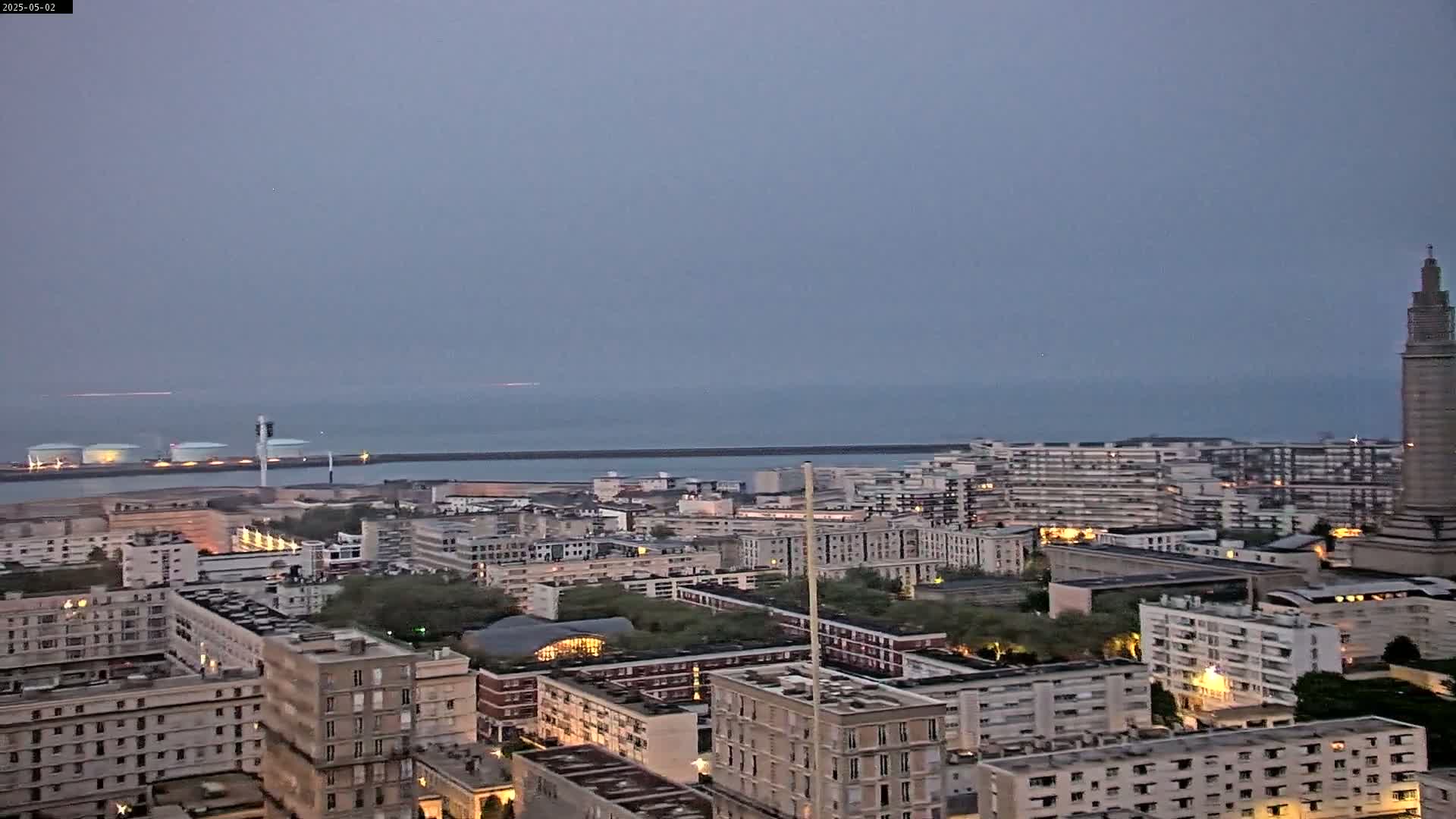 An overcast, twilight view shows a city by the sea, with many apartment buildings and a tall tower in the distance.