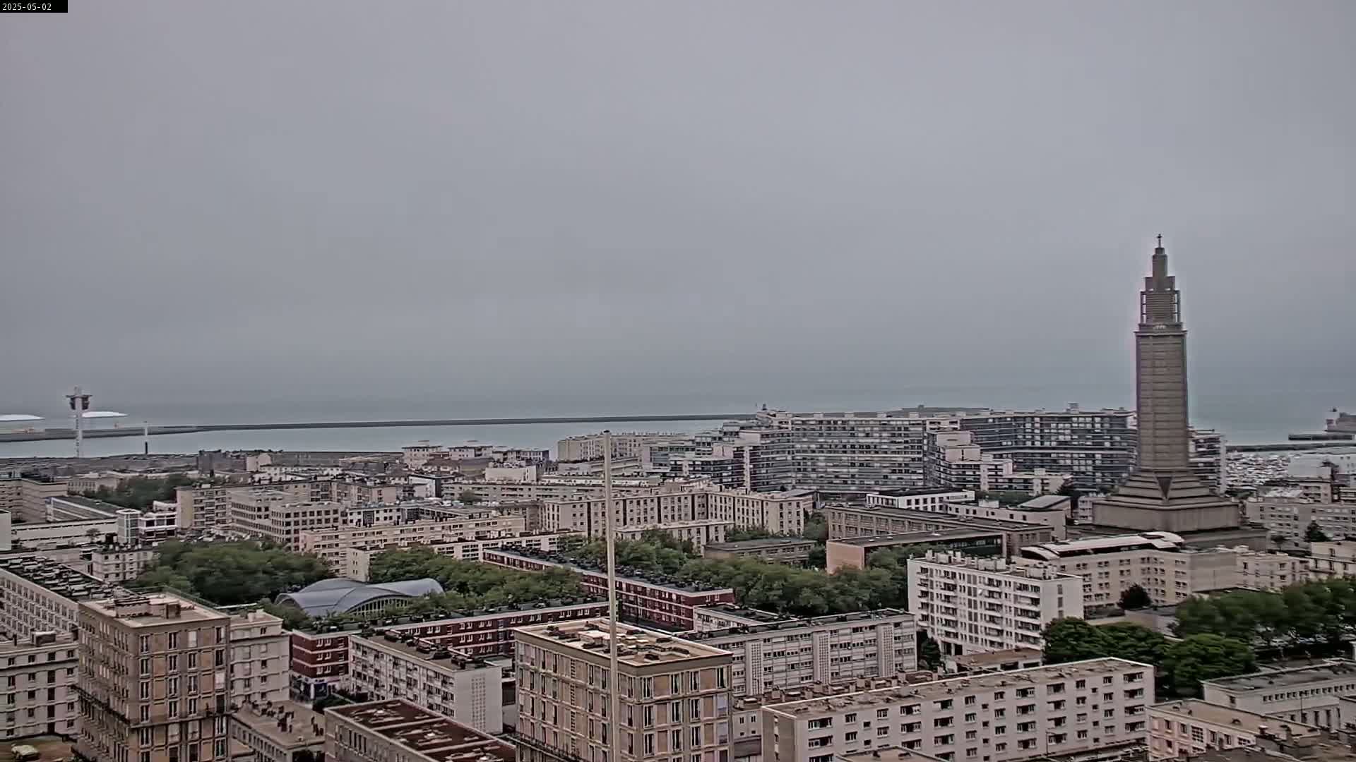 A sprawling cityscape with a tall tower is visible, overlooking a calm, gray sea under an overcast sky.