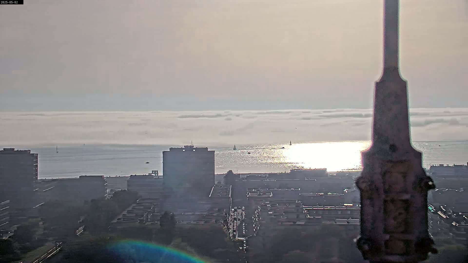 A hazy, sunlit cityscape overlooks a calm body of water covered in a low layer of fog, with several boats visible on the horizon.