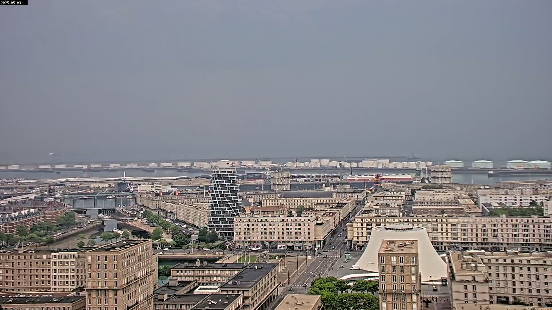 A hazy day reveals a sprawling cityscape with a port featuring numerous industrial storage tanks and ships in the distance, bordered by several multi-story buildings.