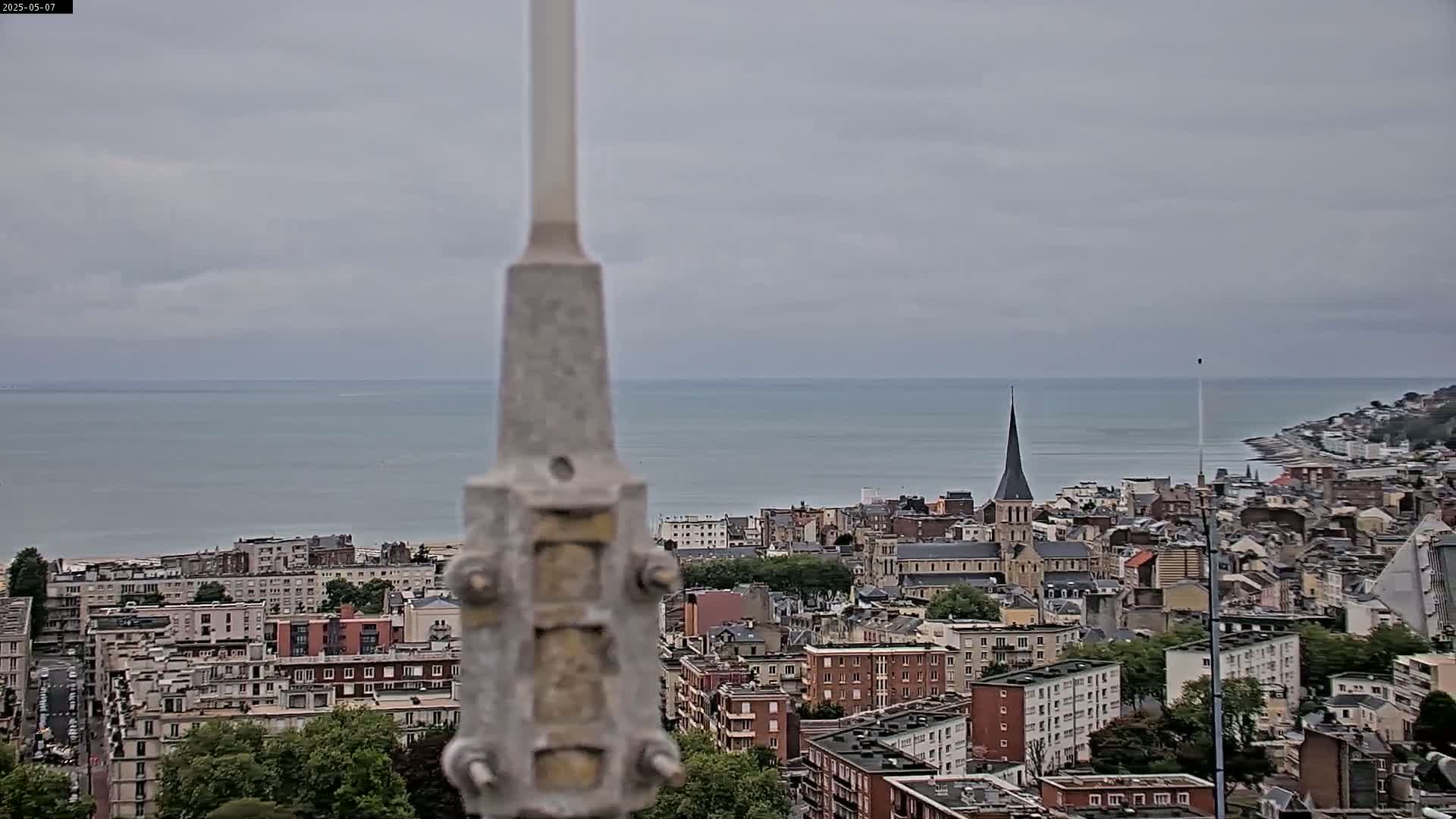 A coastal city with numerous buildings and a church steeple is visible beyond a tall, grey, central structure under an overcast sky.