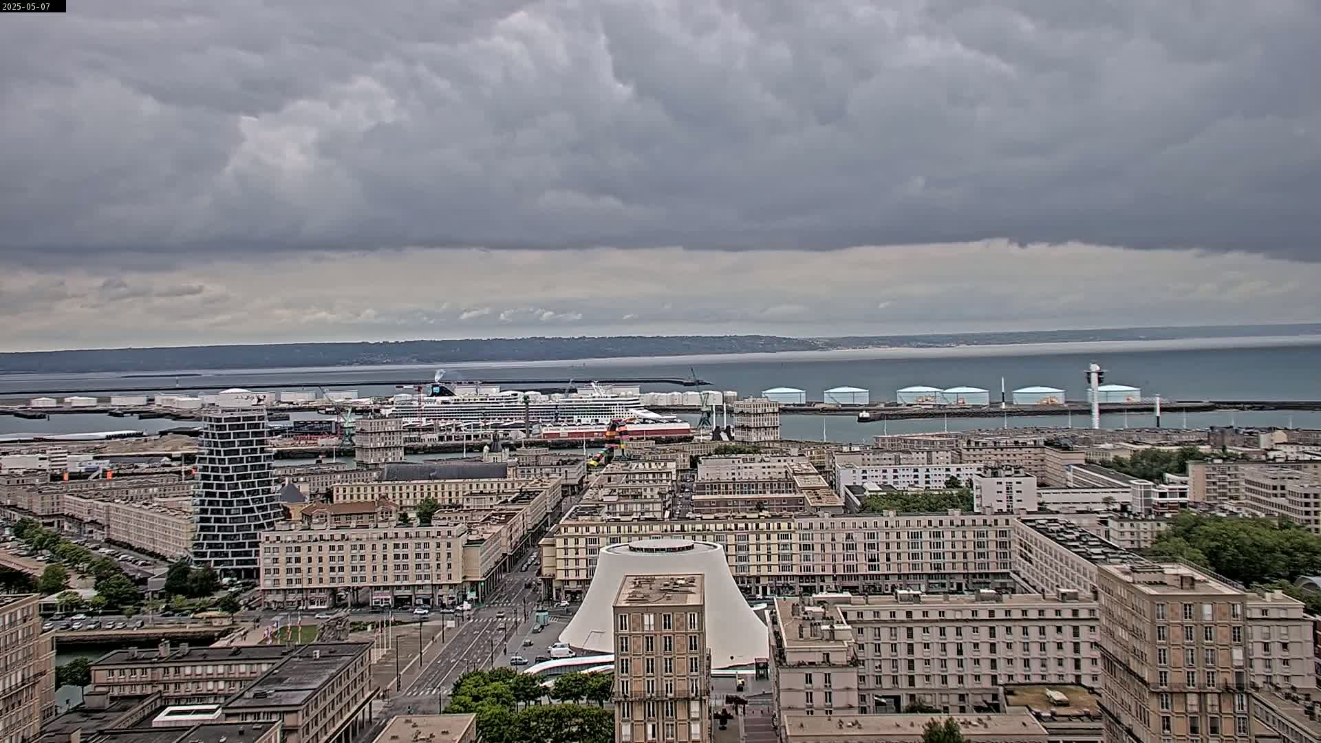 An overcast sky over a city port with several large buildings, a cruise ship, and numerous storage tanks near the water.
