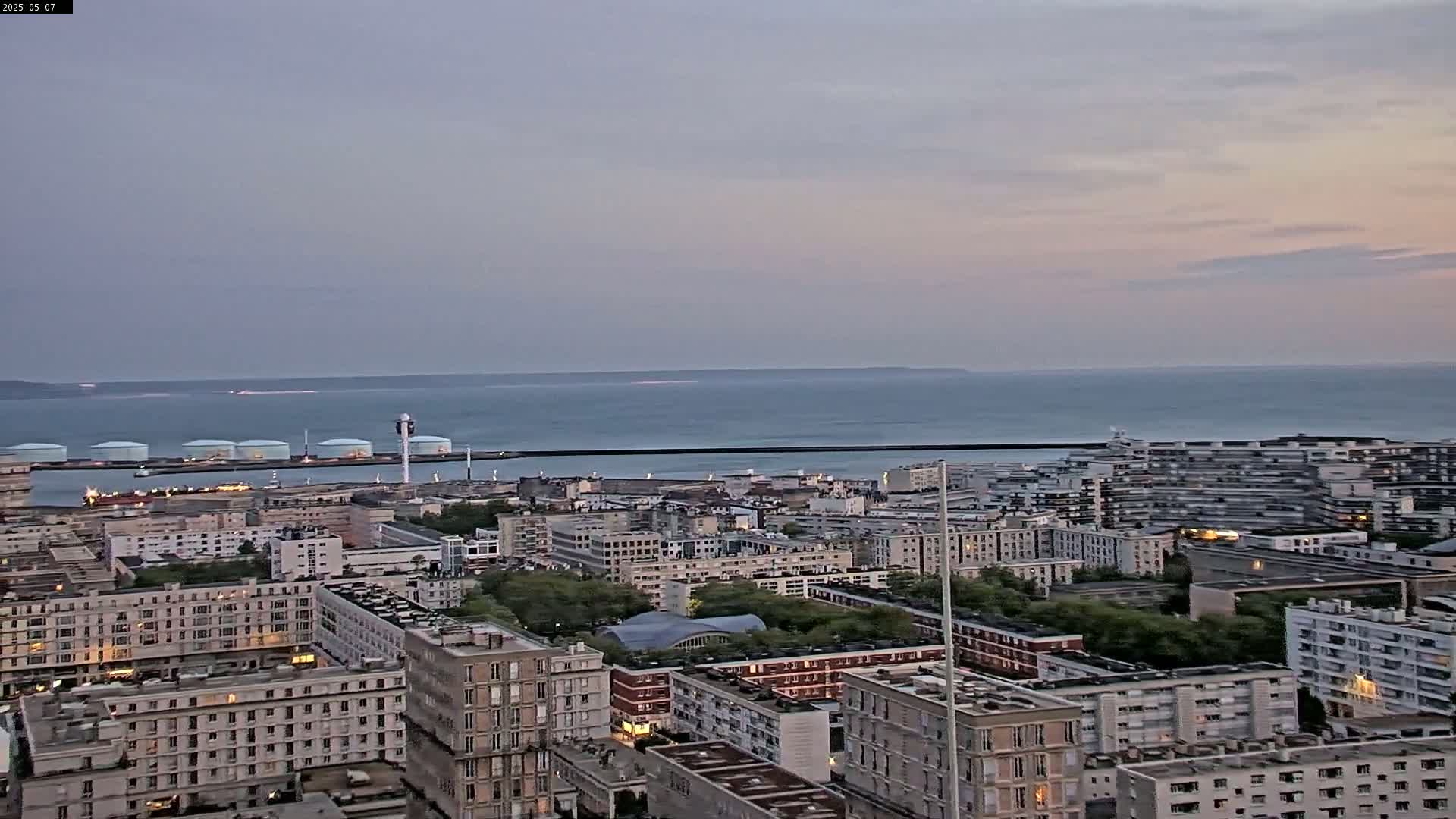 A sprawling city with numerous apartment buildings extends to a harbor with large storage tanks, all under a calm, dusky sky overlooking a tranquil ocean.