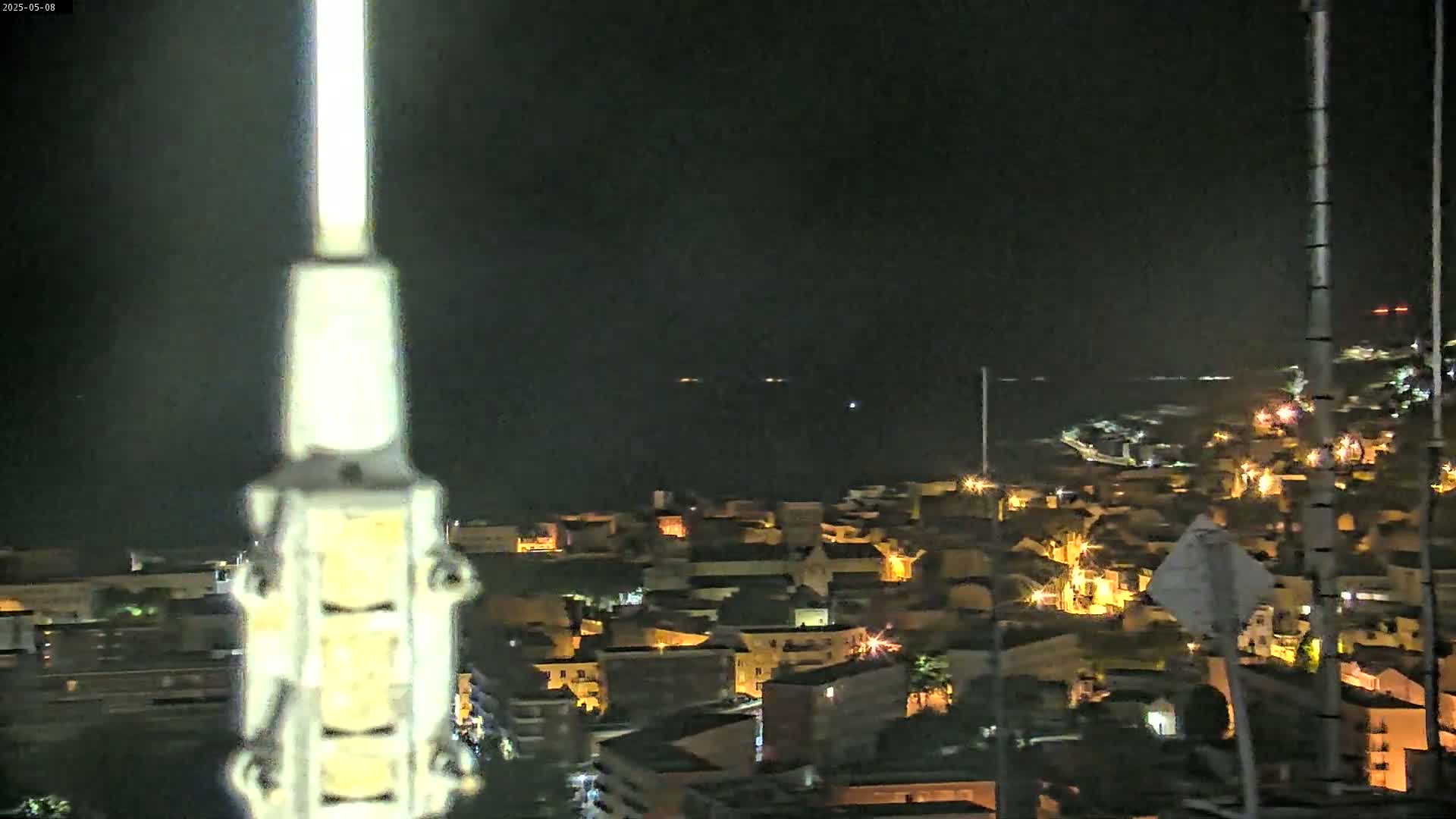 A nighttime view of a coastal town illuminated by streetlights, with a dark body of water visible in the distance under a clear sky.