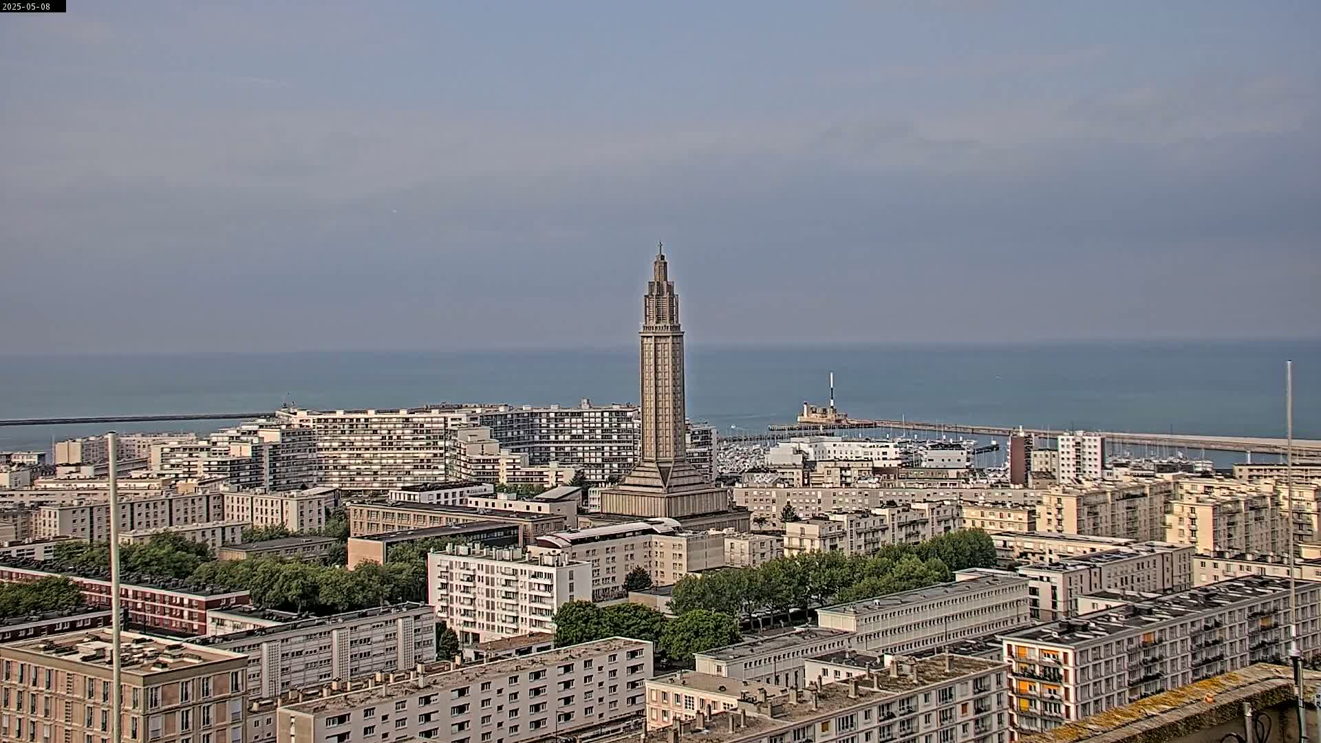 A tall, slender tower stands prominently in a cityscape overlooking a calm ocean under a hazy sky.