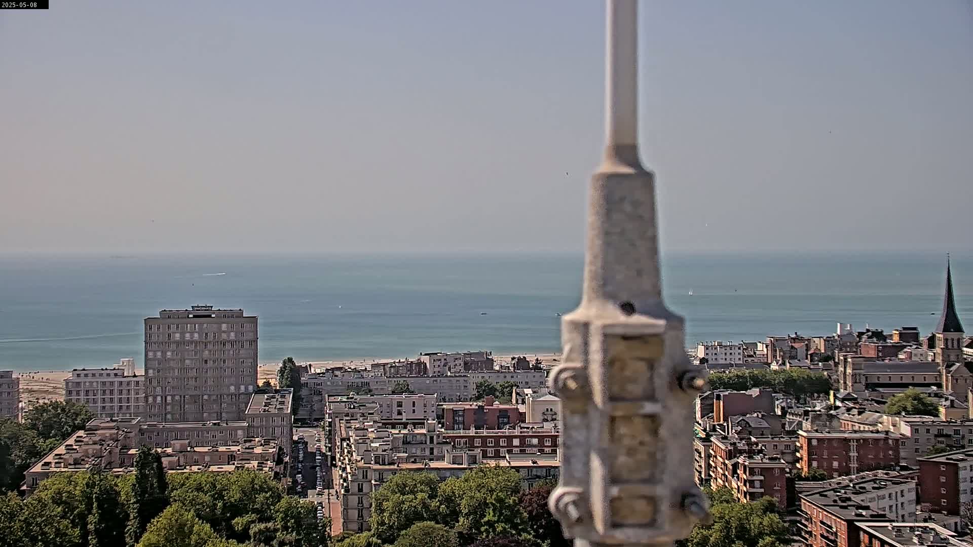 A coastal city scene is visible from a high vantage point on a hazy day, with a calm ocean in the background.