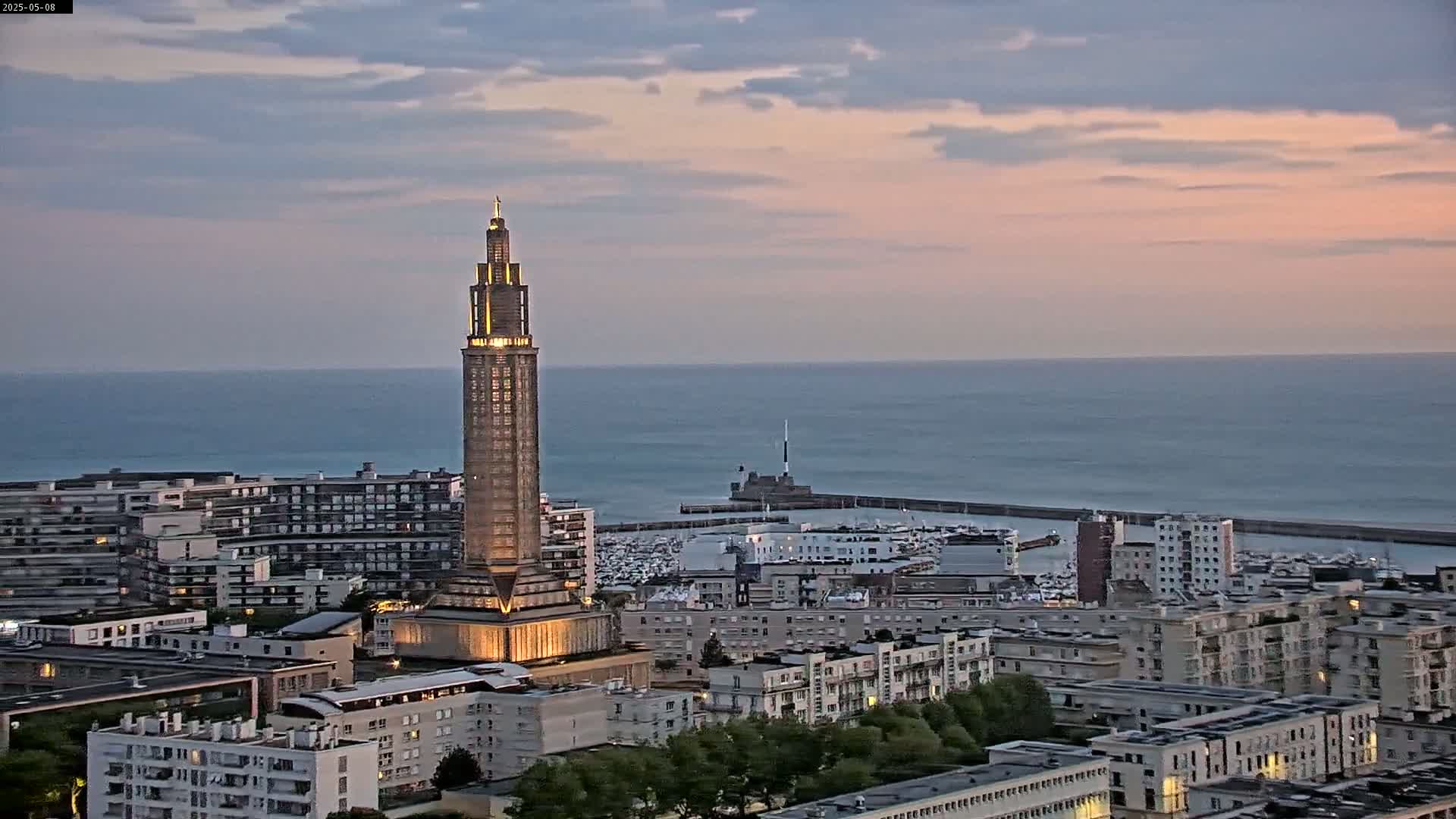 A tall, illuminated tower stands prominently in a coastal city at dusk, overlooking a calm sea under a mostly clear, pinkish sky.