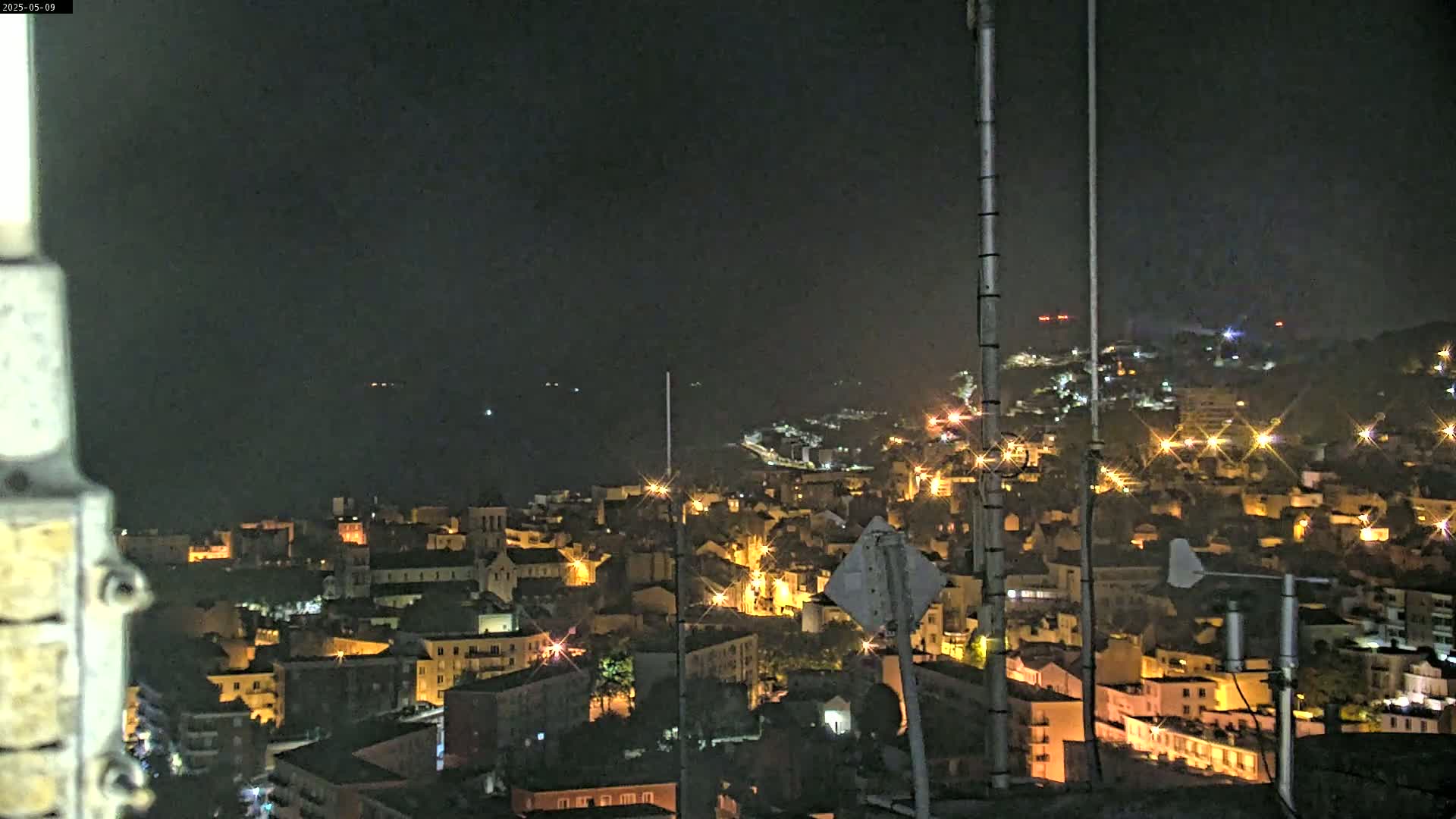 A nighttime view of a coastal town illuminated by streetlights, with a dark sea in the background under a clear sky.