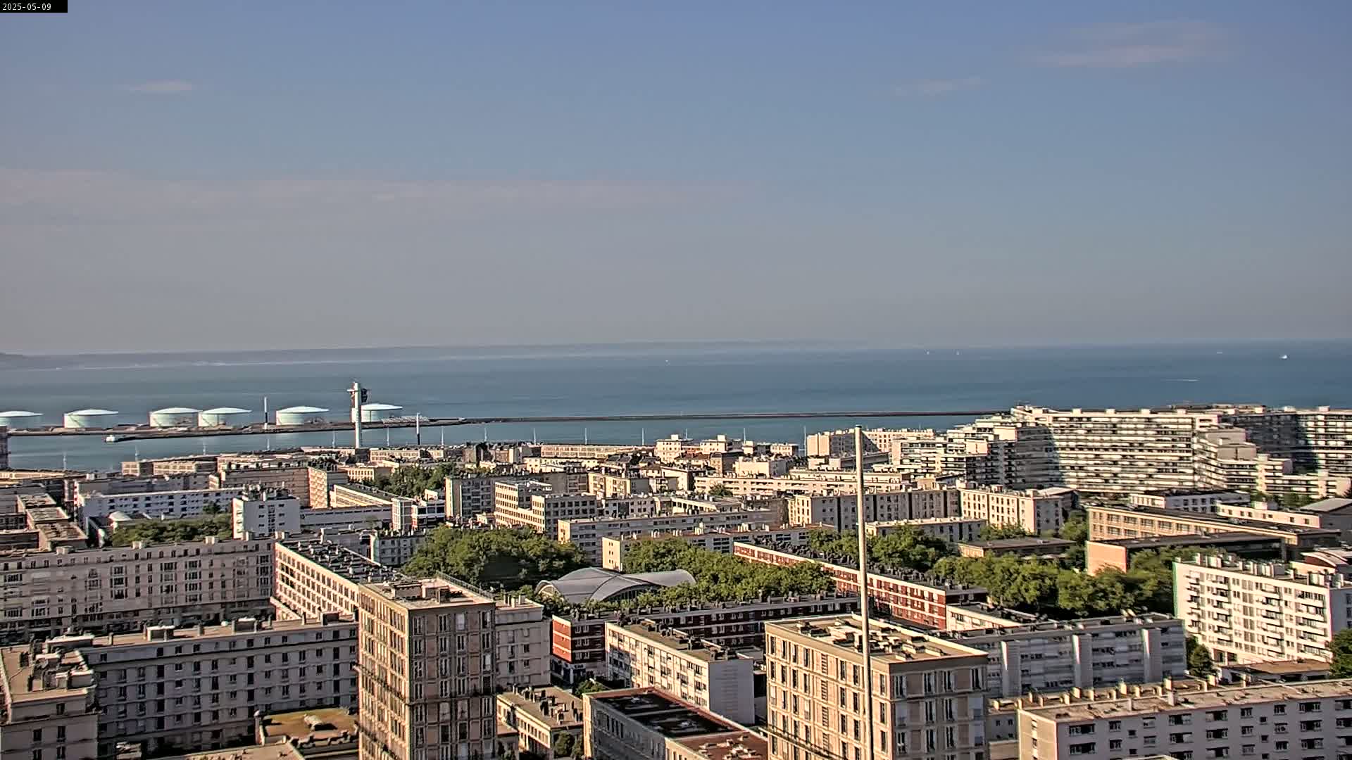 A sprawling cityscape of light-colored apartment buildings extends to a calm, blue ocean under a mostly clear sky.