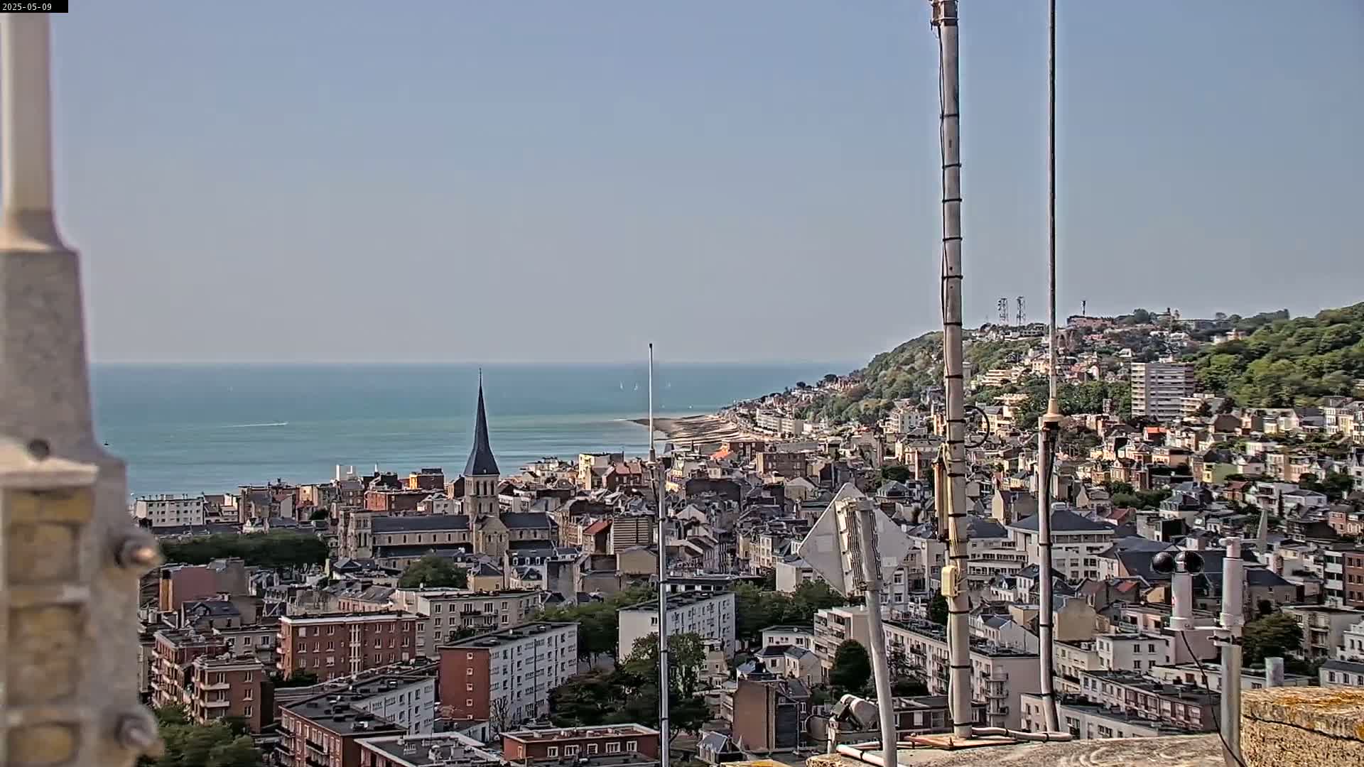A coastal city with a church spire is visible from a high vantage point on a clear day, with a calm ocean in the background.