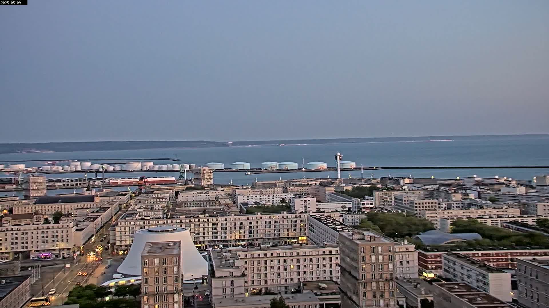 A high-angle, long shot of a city with a port and calm ocean under a clear, twilight sky.