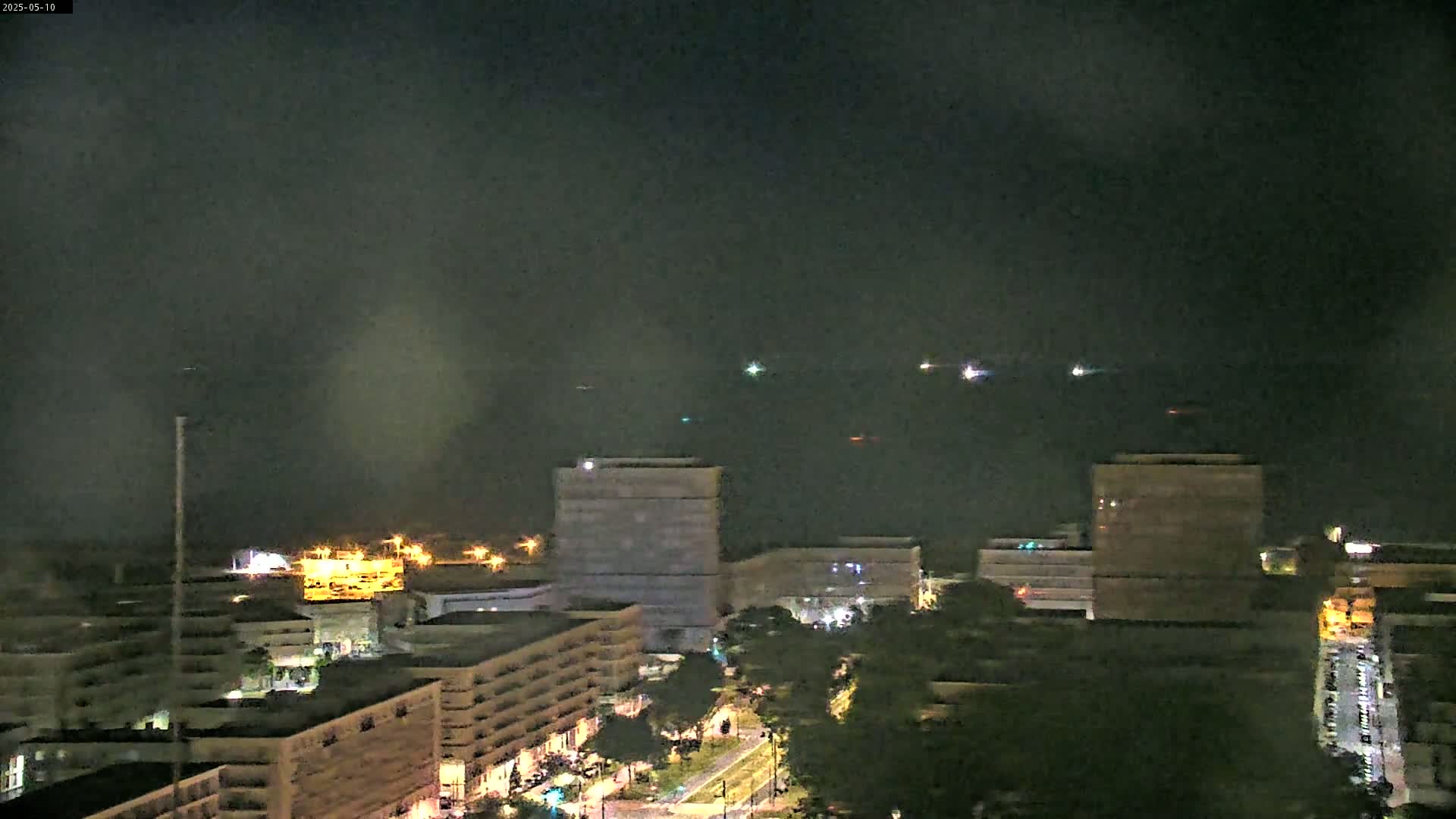 A nighttime view of city buildings and a body of water with faint lights on it, under a hazy, dark sky.