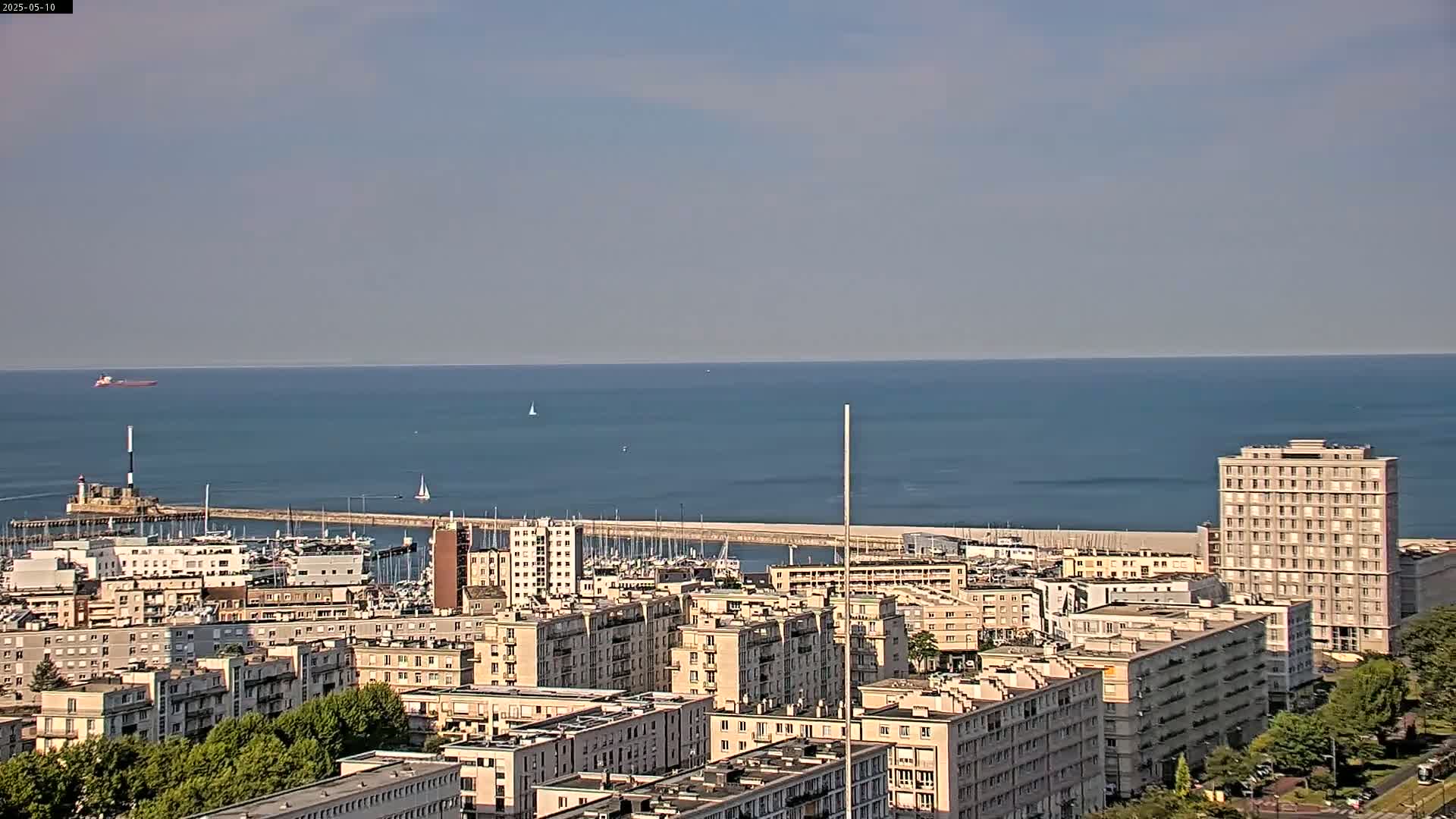 A coastal city with a marina and numerous apartment buildings is seen on a clear, sunny day, overlooking a calm ocean with a distant cargo ship.