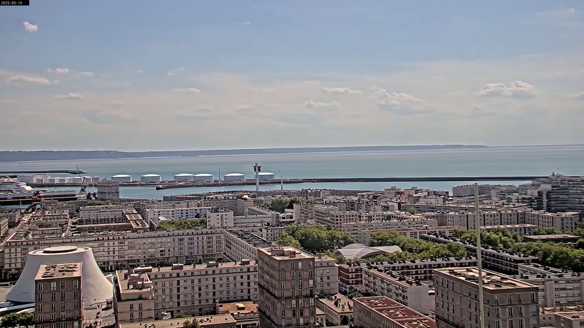 A coastal city with a harbor filled with ships and storage tanks, under a partly cloudy sky.