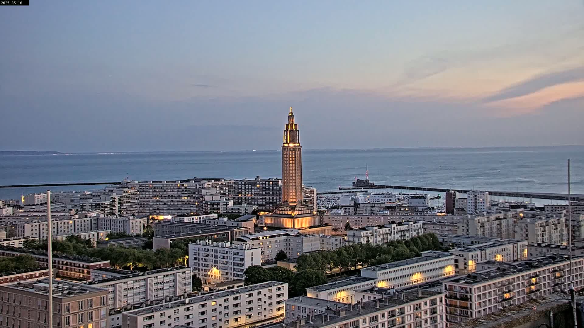 A tall, illuminated tower stands in the center of a coastal city at dusk, overlooking a calm sea.