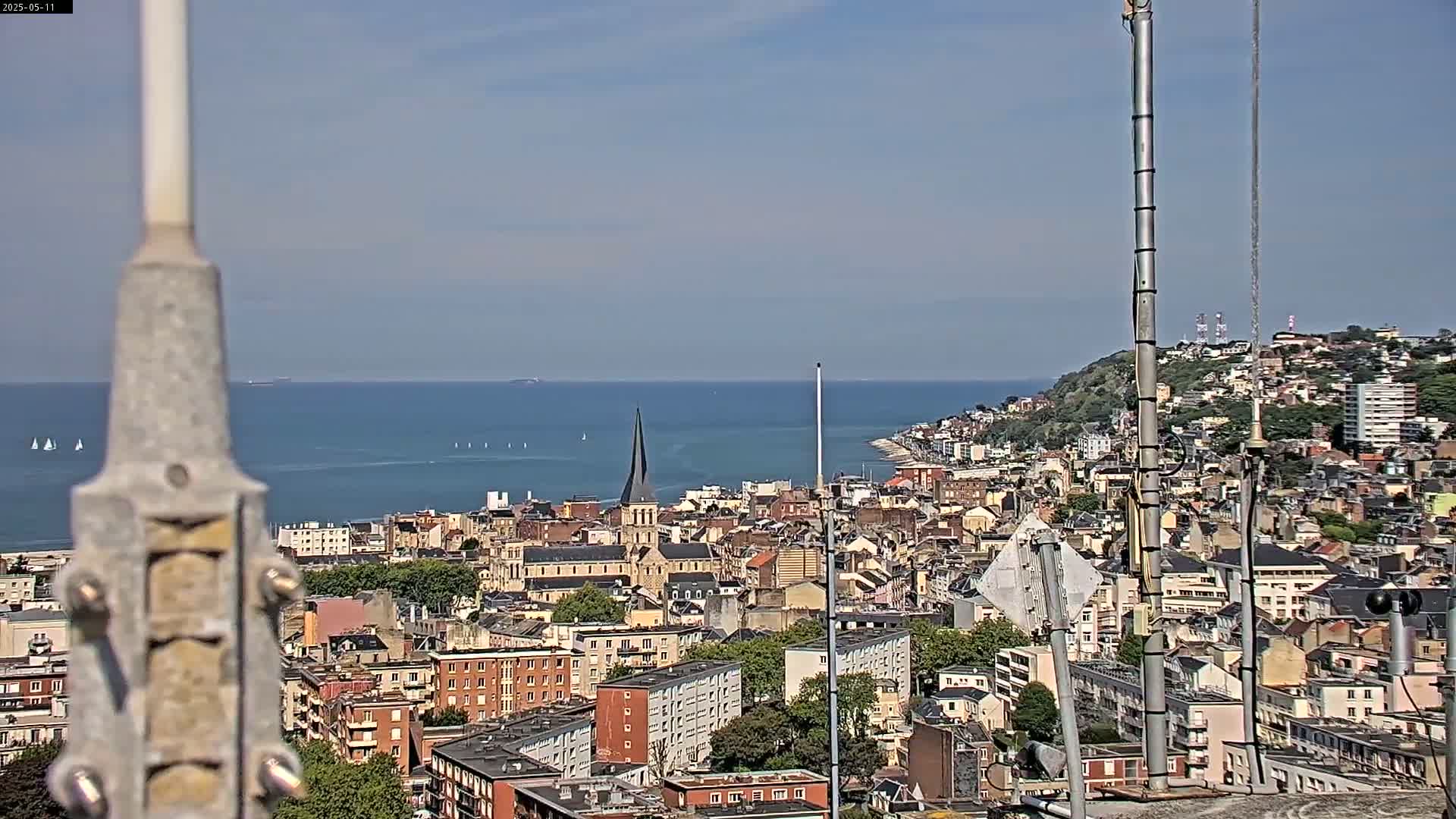 A coastal city with numerous buildings is visible from a high vantage point on a clear, sunny day, with a body of water and sailboats in the distance.