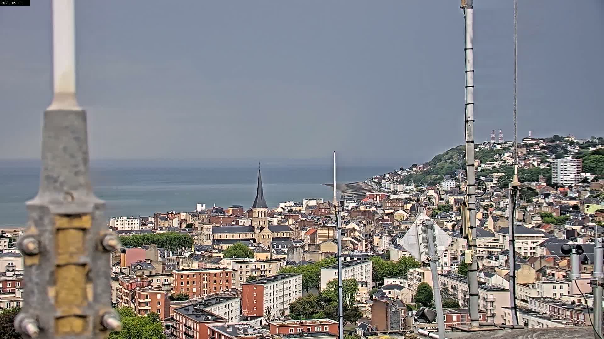 A coastal city with a church steeple is seen on an overcast day, viewed from a high vantage point near several antenna towers.