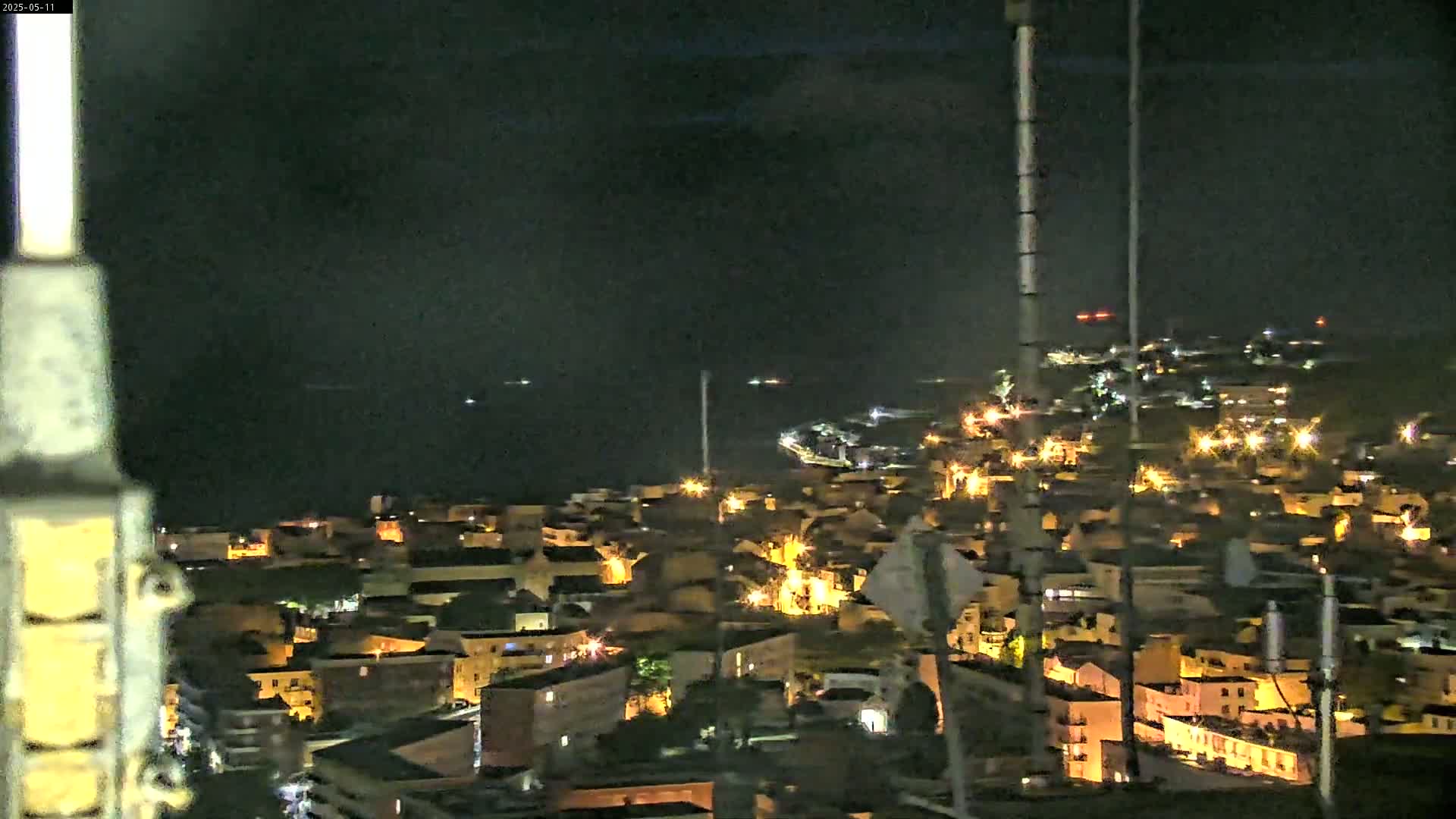 A nighttime, high-angle view shows a coastal town illuminated with scattered lights, overlooking a dark body of water under a clear, dark sky.