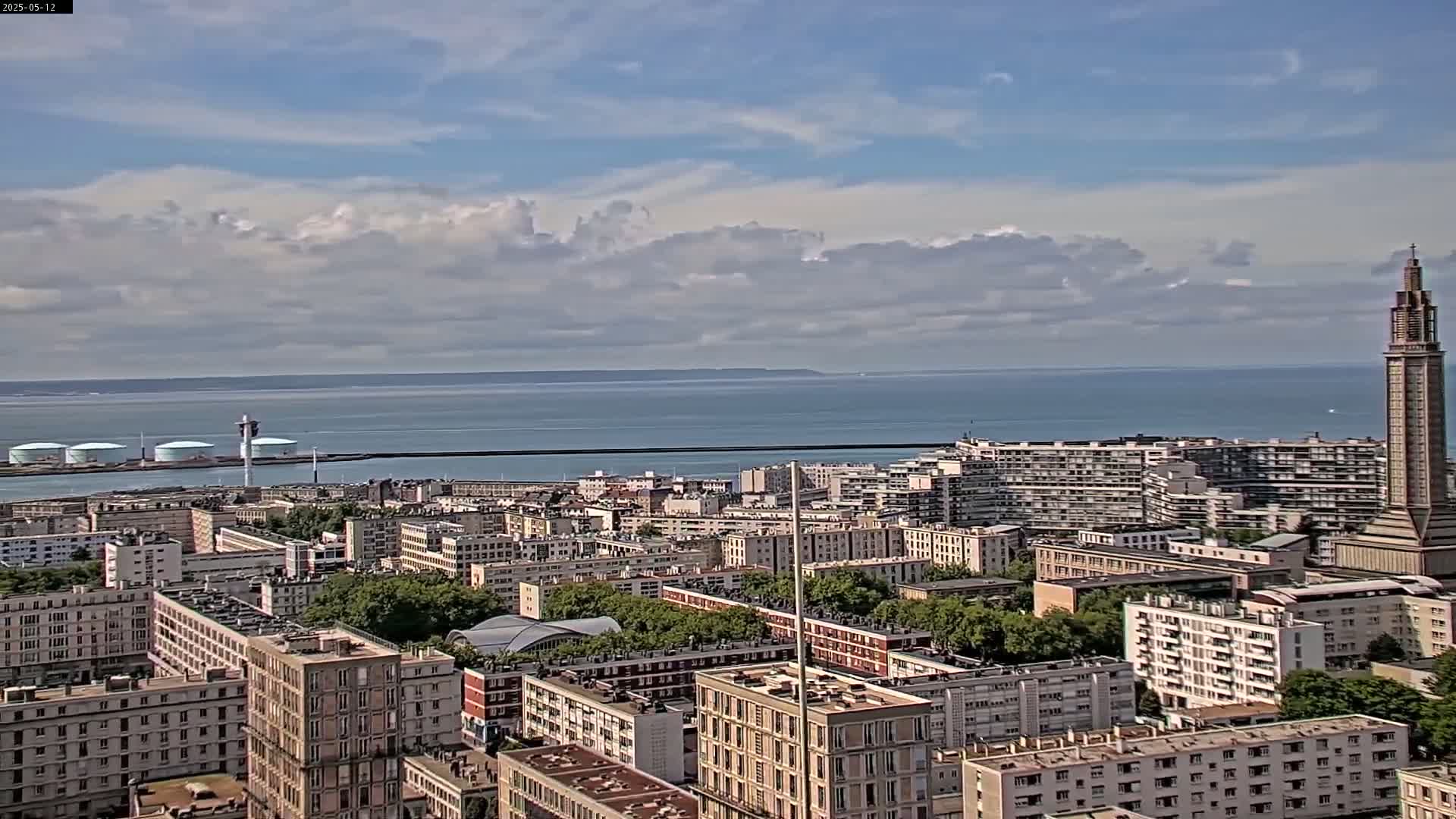 A coastal city with numerous apartment buildings stretches toward a body of water under a partly cloudy sky.