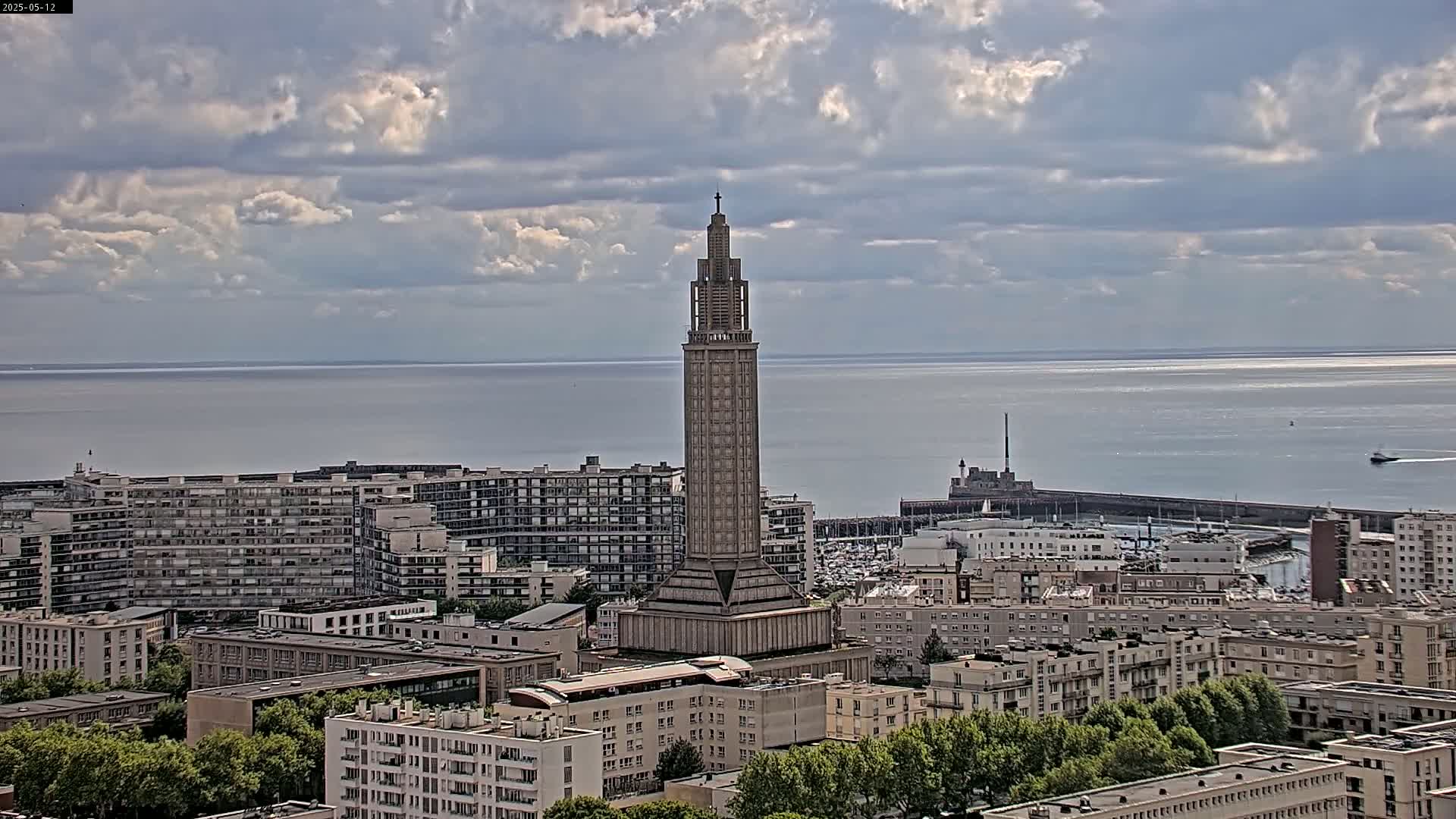 A tall, slender tower stands in the center of a cityscape overlooking a calm ocean under a partly cloudy sky.