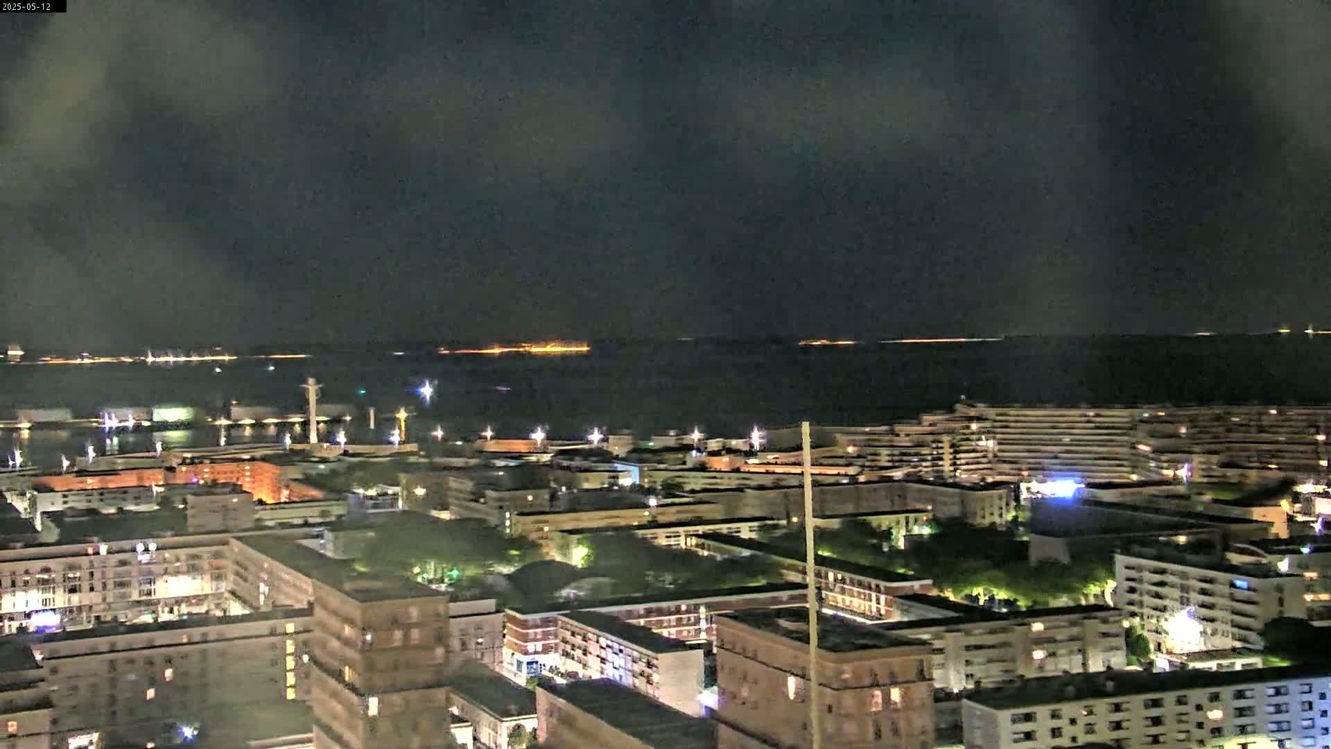 A nighttime aerial view shows a city's waterfront illuminated, with a dark, cloudy sky above and a body of water extending to a distant, faintly lit horizon.