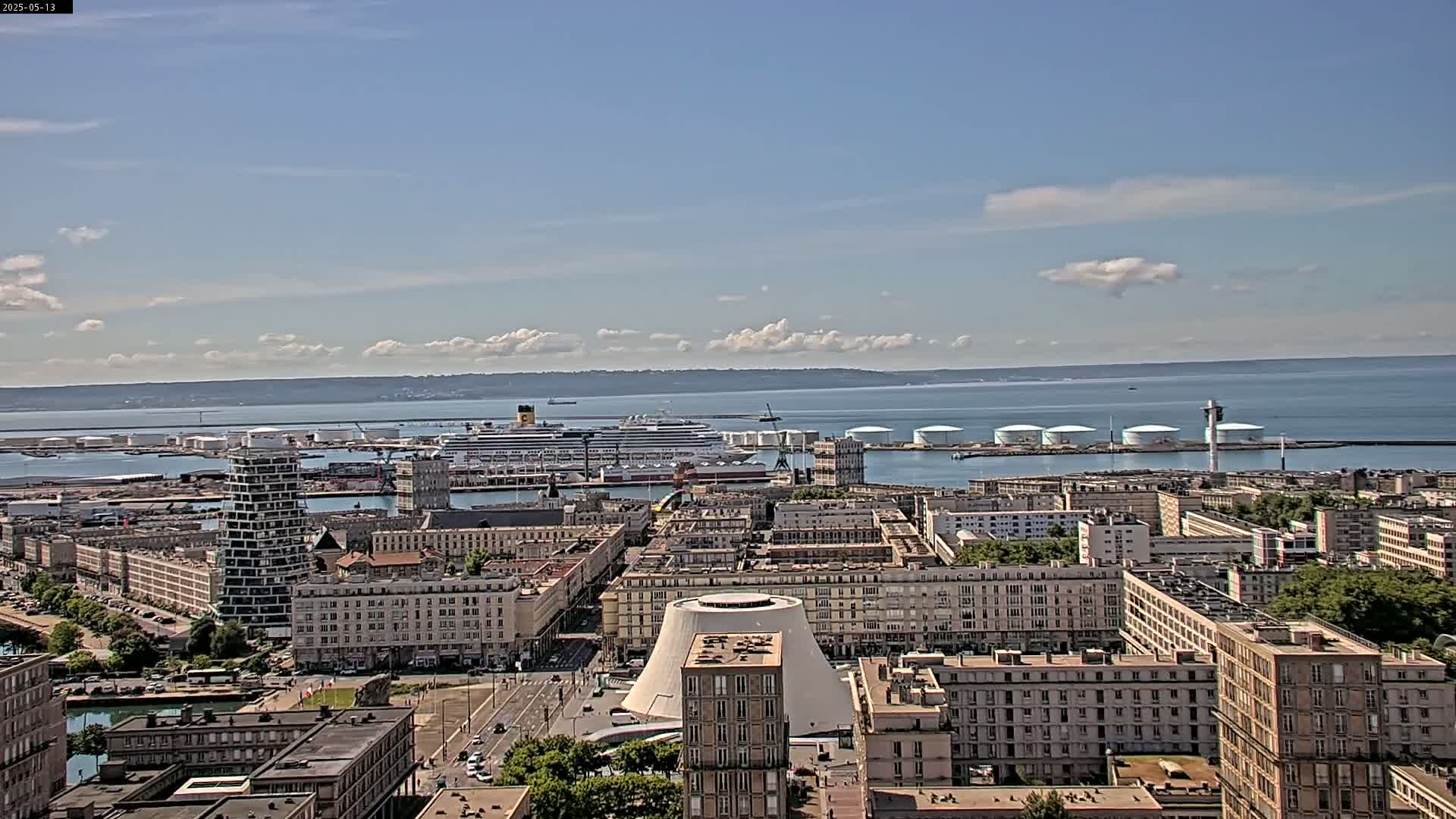 A large cruise ship is docked in a harbor city under a mostly sunny sky with some scattered clouds; the city extends inland from the water, showcasing a mix of building styles and heights.