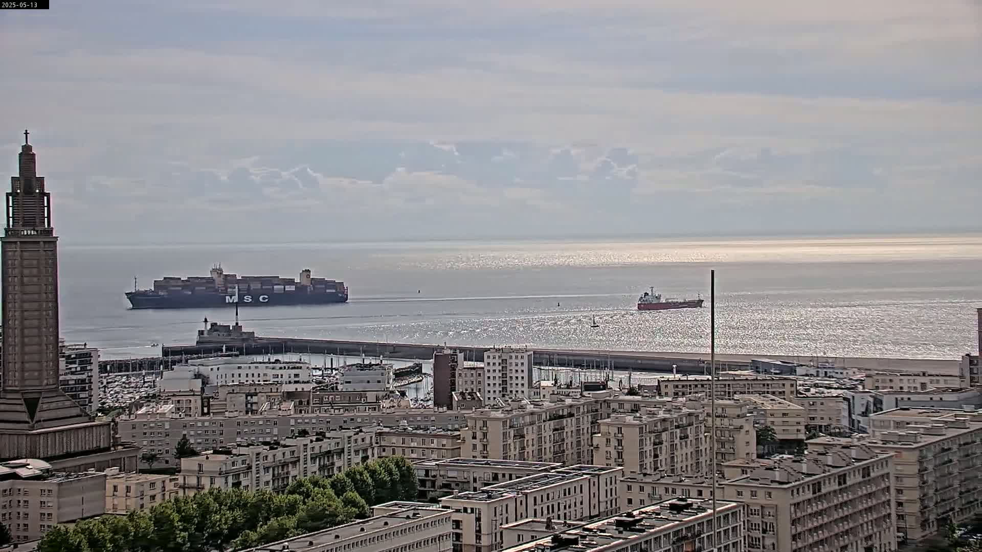 A large container ship is visible in the harbor, with a smaller ship further out at sea, under a partly cloudy sky.
