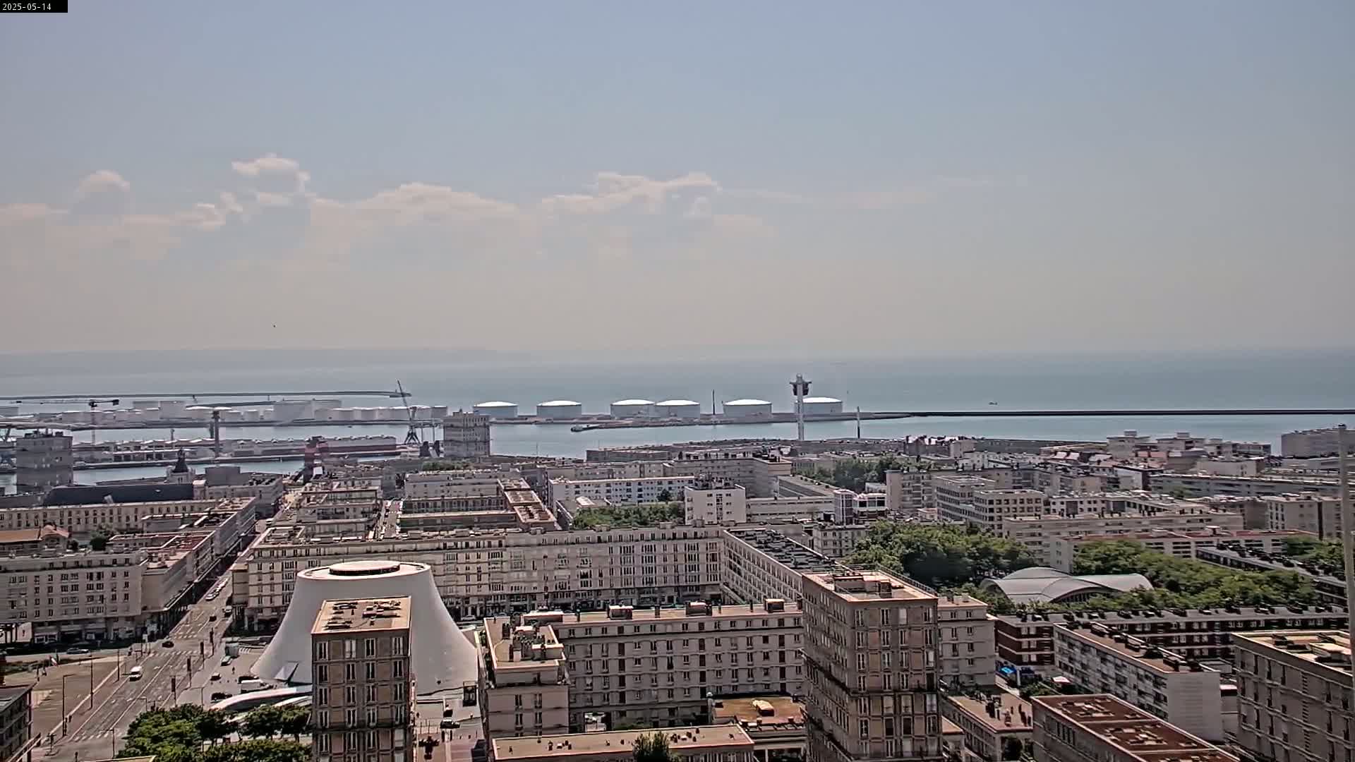 A partly cloudy day reveals a cityscape next to a harbor with several large storage tanks, and a calm sea extending to the horizon.