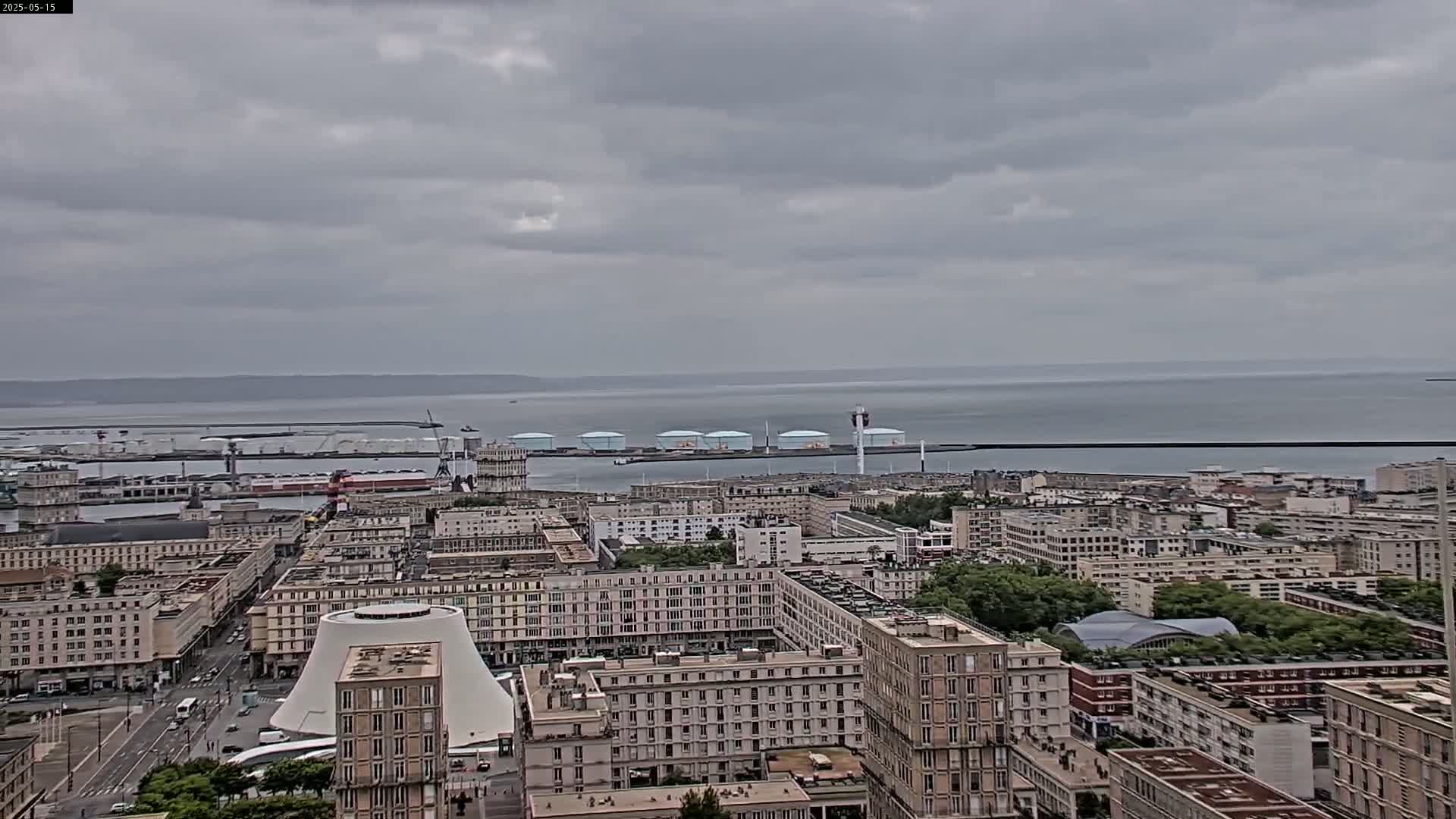 A coastal city with numerous buildings is visible under an overcast sky, with a body of water and industrial structures in the background.