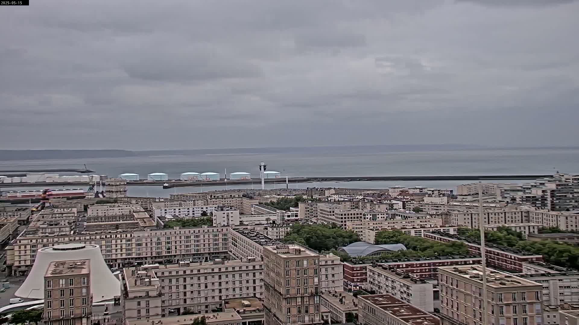 A coastal city with numerous buildings under an overcast sky overlooks a calm body of water.