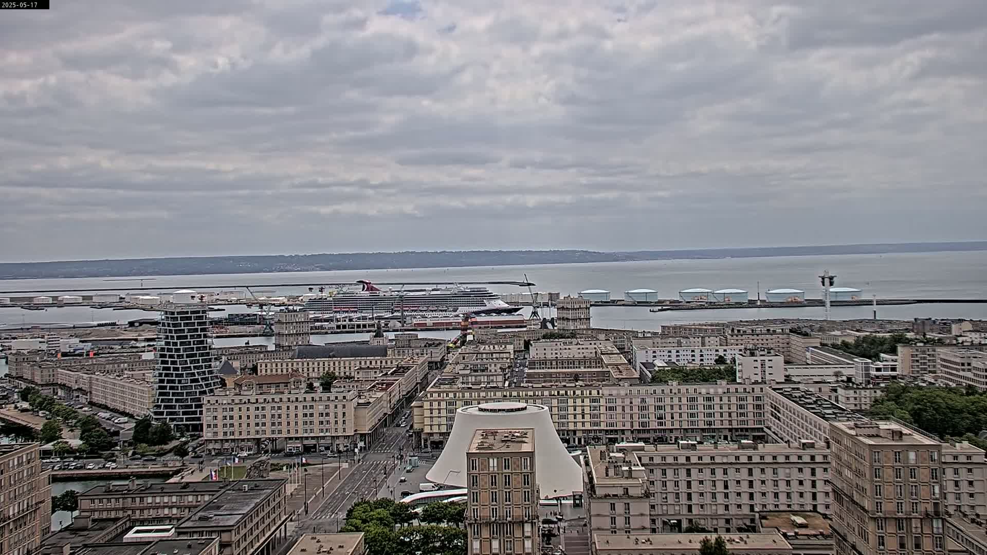 An overcast day reveals a city harbor with a large cruise ship docked, and numerous buildings extending to a calm body of water under a cloudy sky.