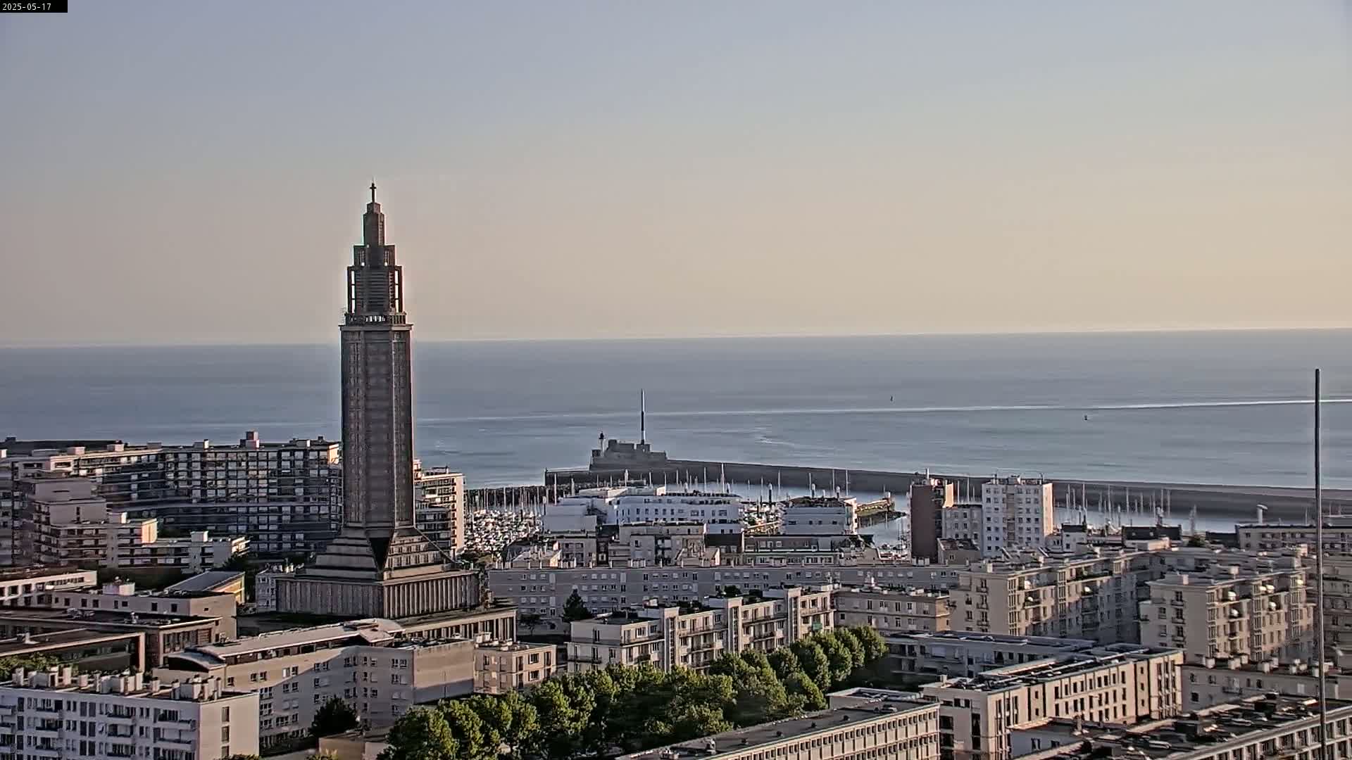 A tall, slender tower stands prominently in a cityscape overlooking a calm ocean under a clear sky.