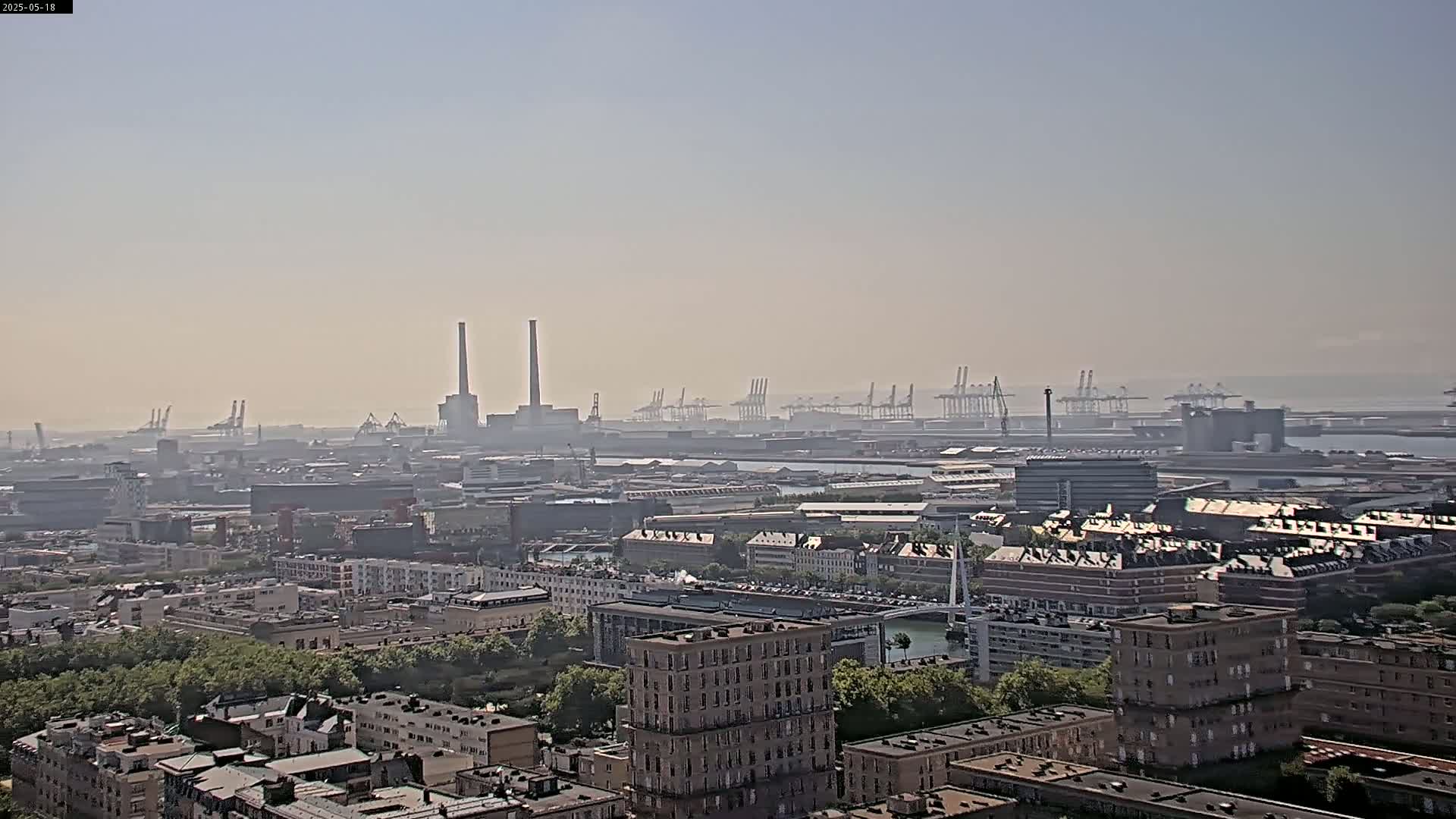 A hazy, distant view of an industrial port area with numerous cranes and smokestacks, overlooked by a cityscape with buildings and greenery.