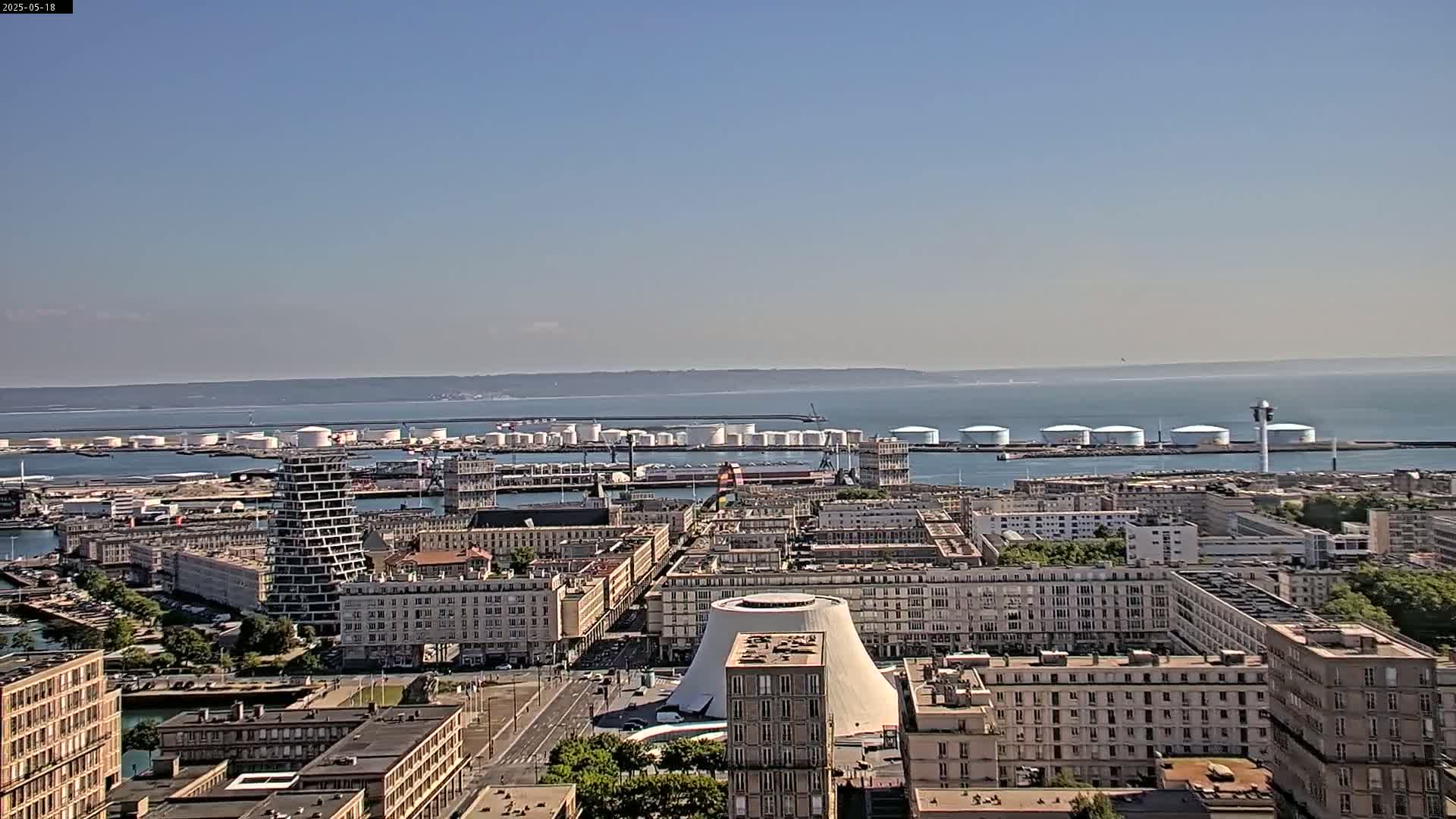 A high-angle, long shot on a clear, sunny day shows a city's harbor with numerous large, white storage tanks, a cityscape of multi-story buildings, and a body of water extending to the horizon.
